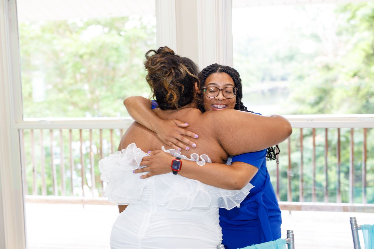 Two women sharing a warm embrace at a bright indoor venue, one wearing a white dress and the other in royal blue.