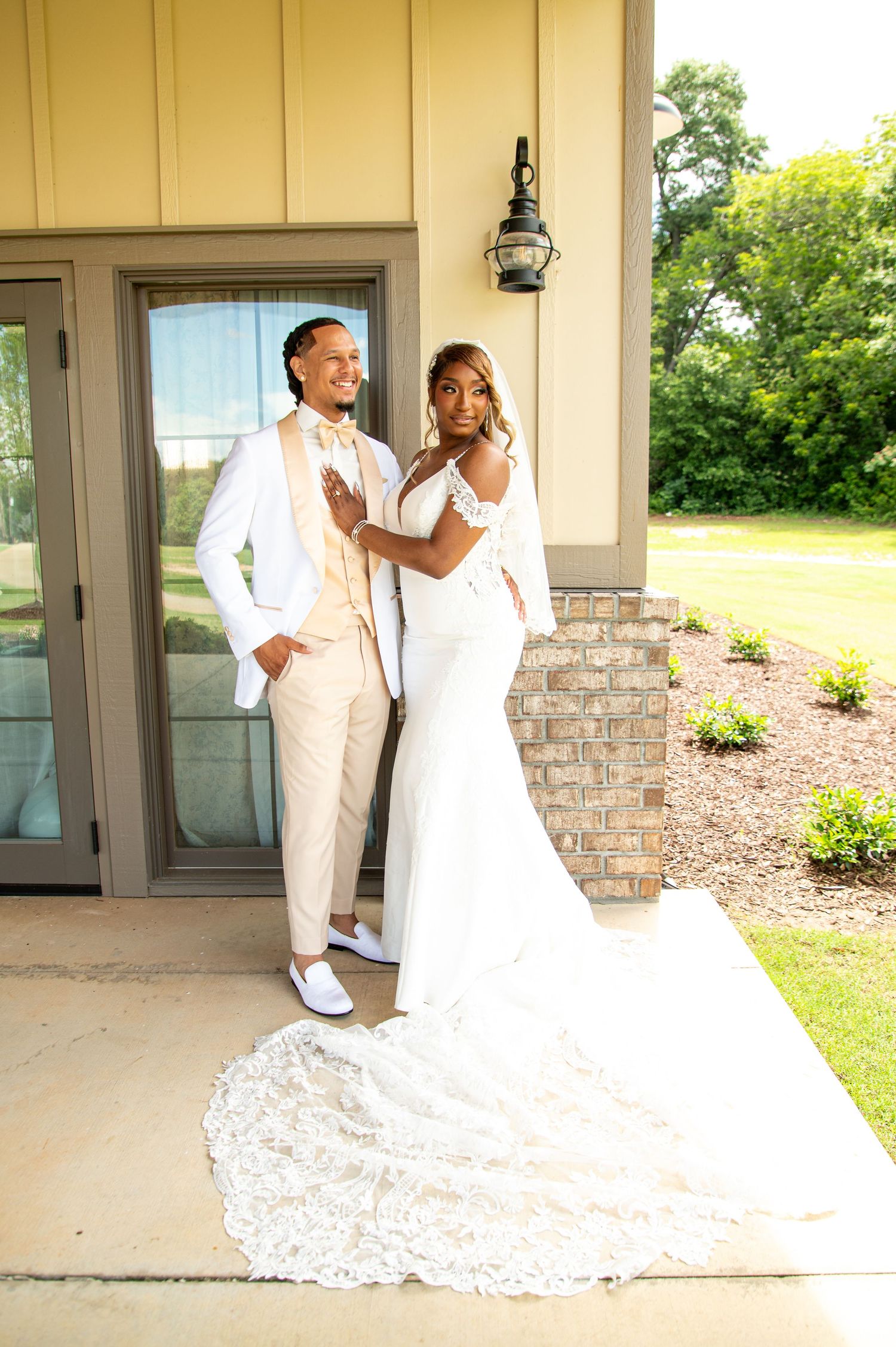 Bride and groom pose together outside venue, bride in lace gown with long train.