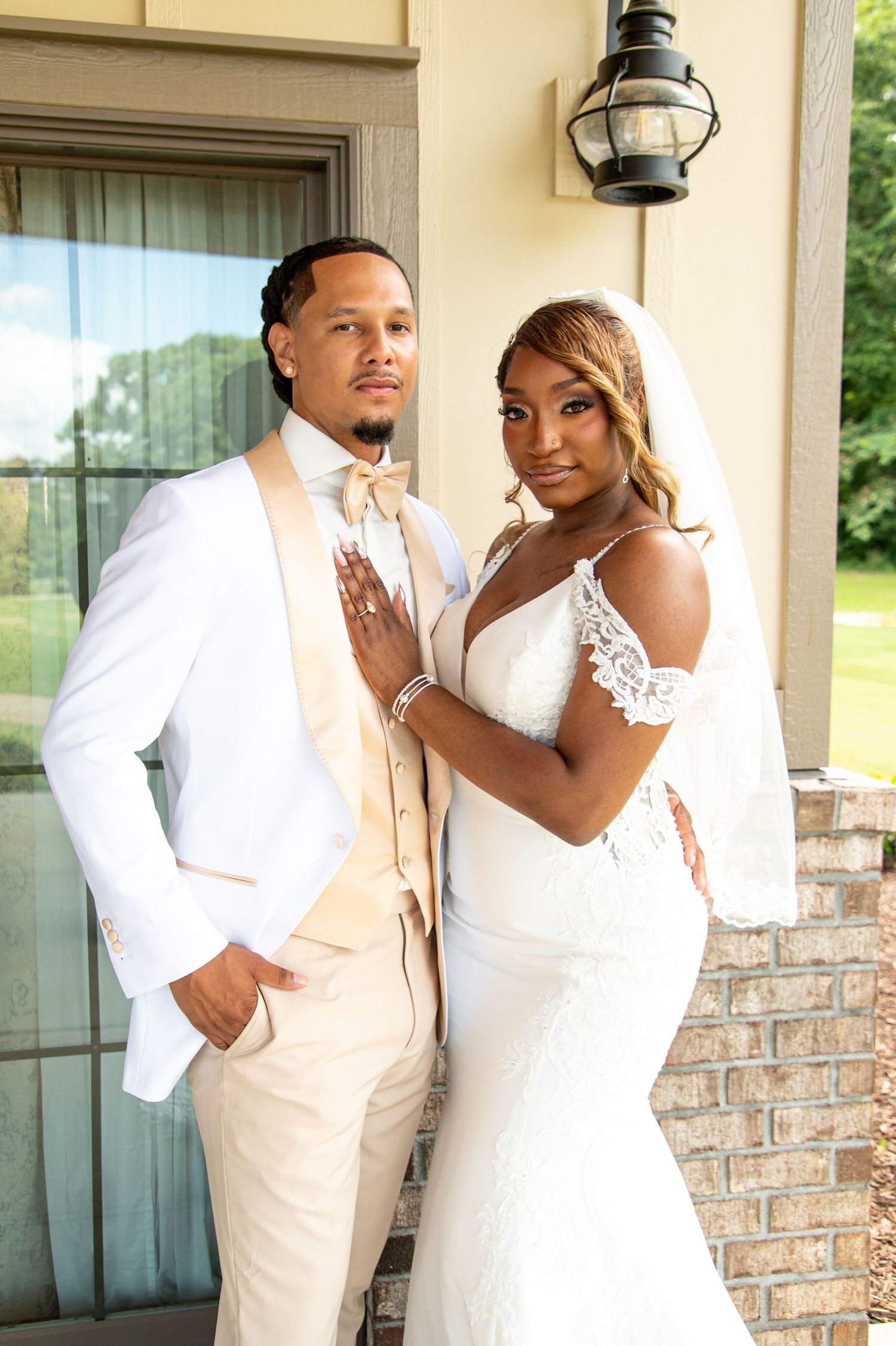 Elegant bride and groom posing together on wedding day, bride in lace mermaid gown with veil, groom in white tuxedo.
