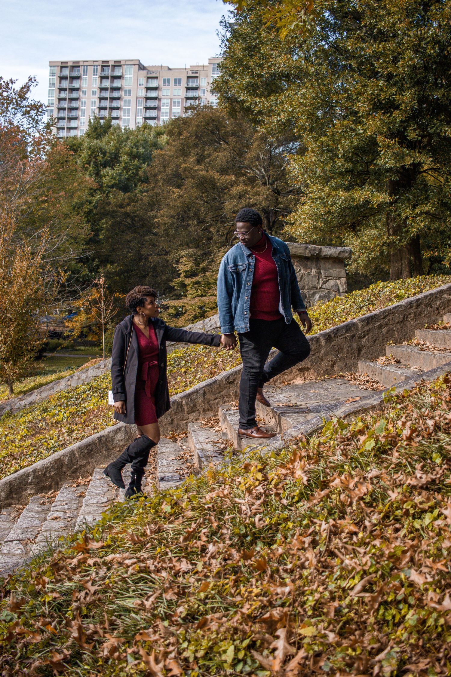Person in blue denim jacket and red sweater walks up stone steps surrounded by autumn leaves in urban park setting.