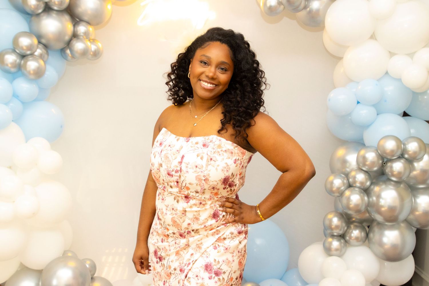 Person in a floral dress posing against a backdrop of blue and silver balloon decorations.