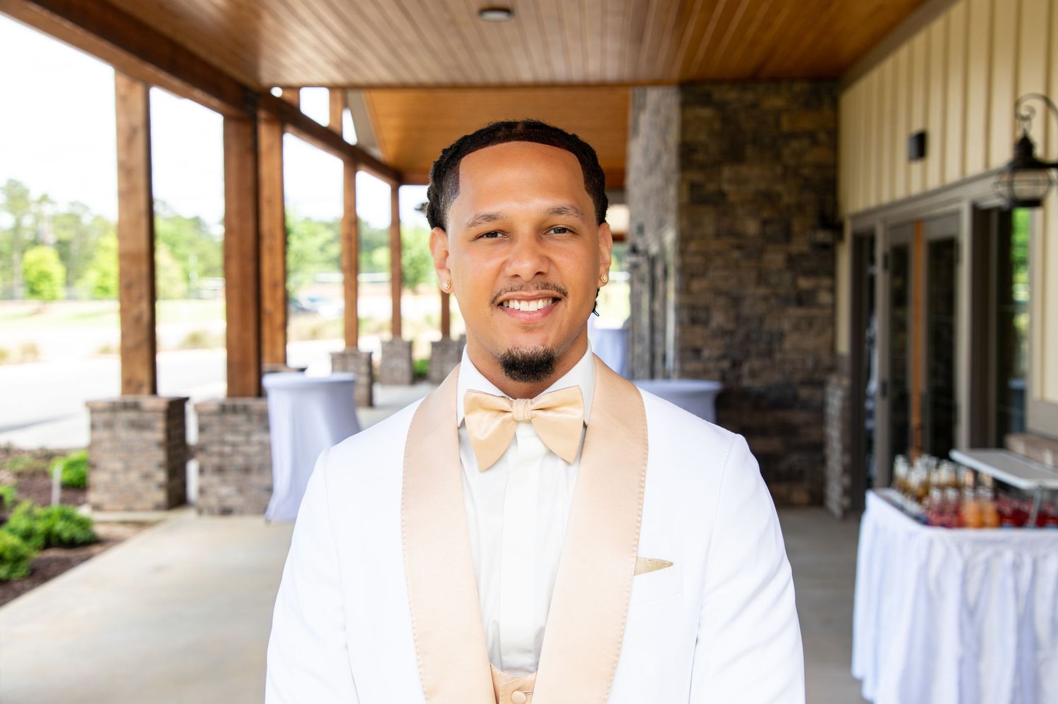 Smiling groom in white jacket with champagne bow tie standing outside elegant wedding venue entrance.