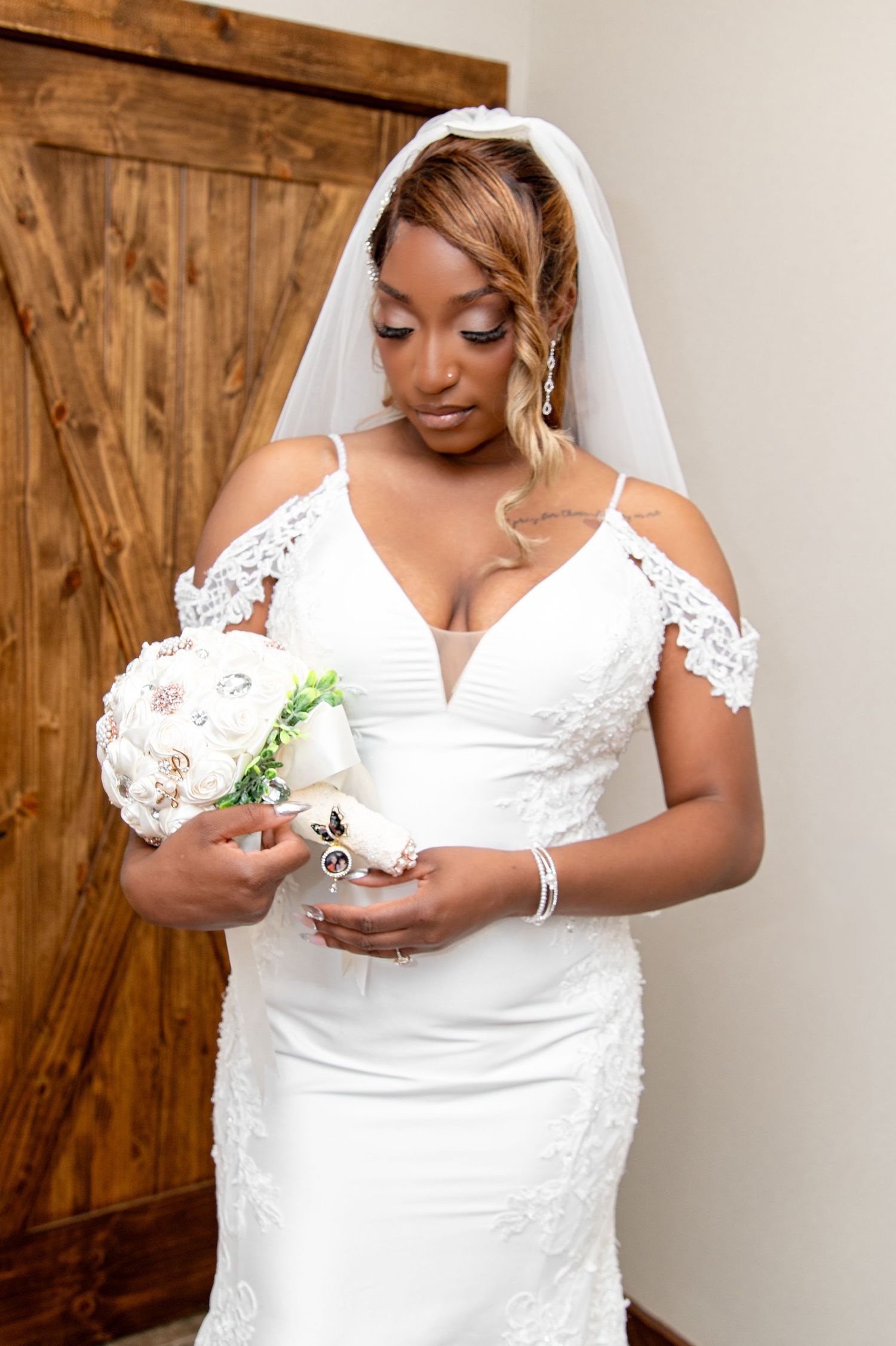 Elegant bride in a white lace off-shoulder wedding gown holding a bouquet near a barn door.