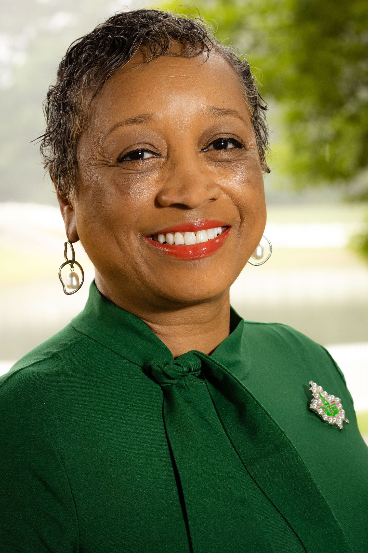 Smiling woman in green blouse with brooch and hoop earrings, posing outdoors with blurred green trees background.