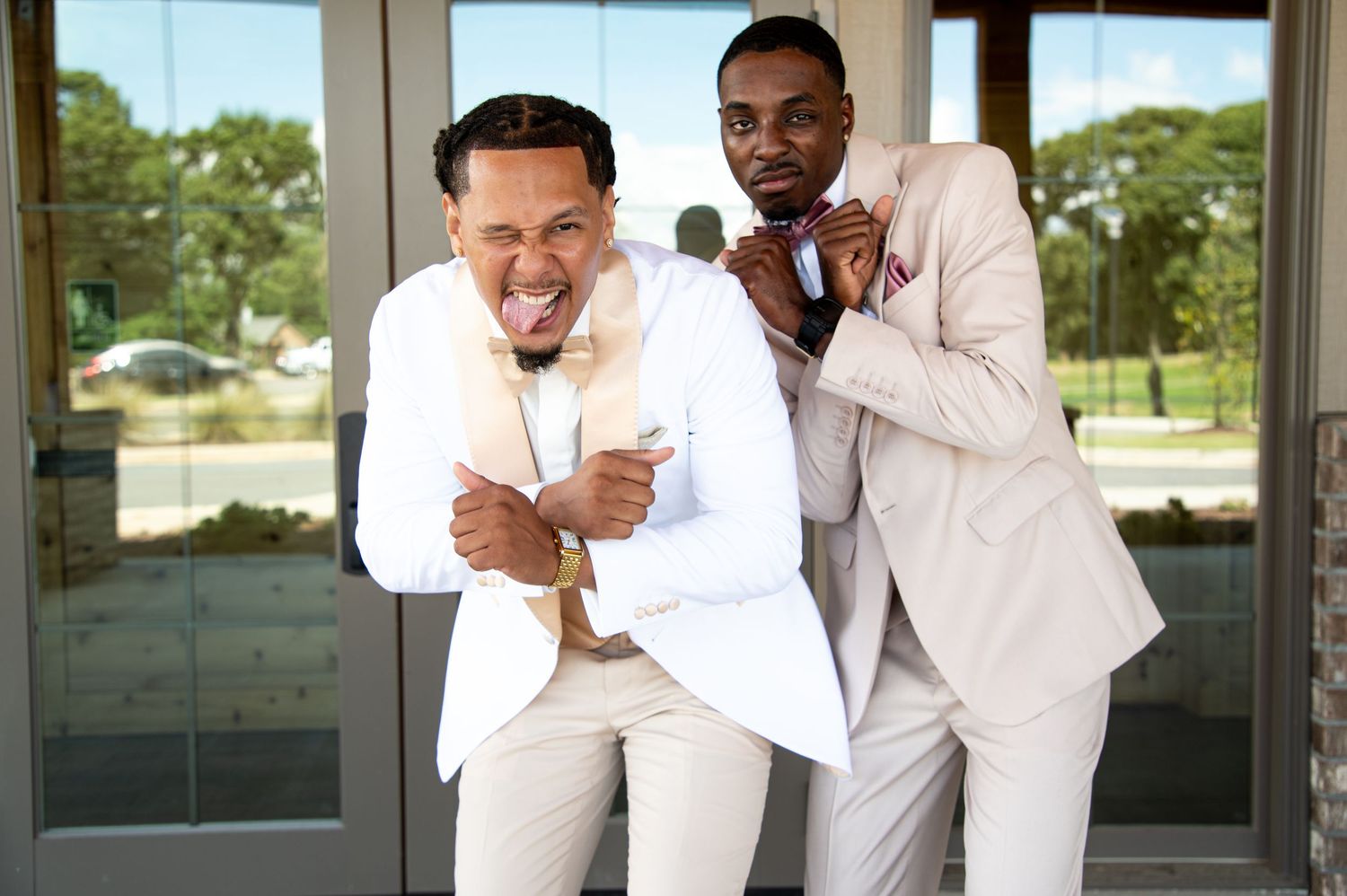 Two groomsmen in cream and white suits laughing and posing together outside wedding venue doors.