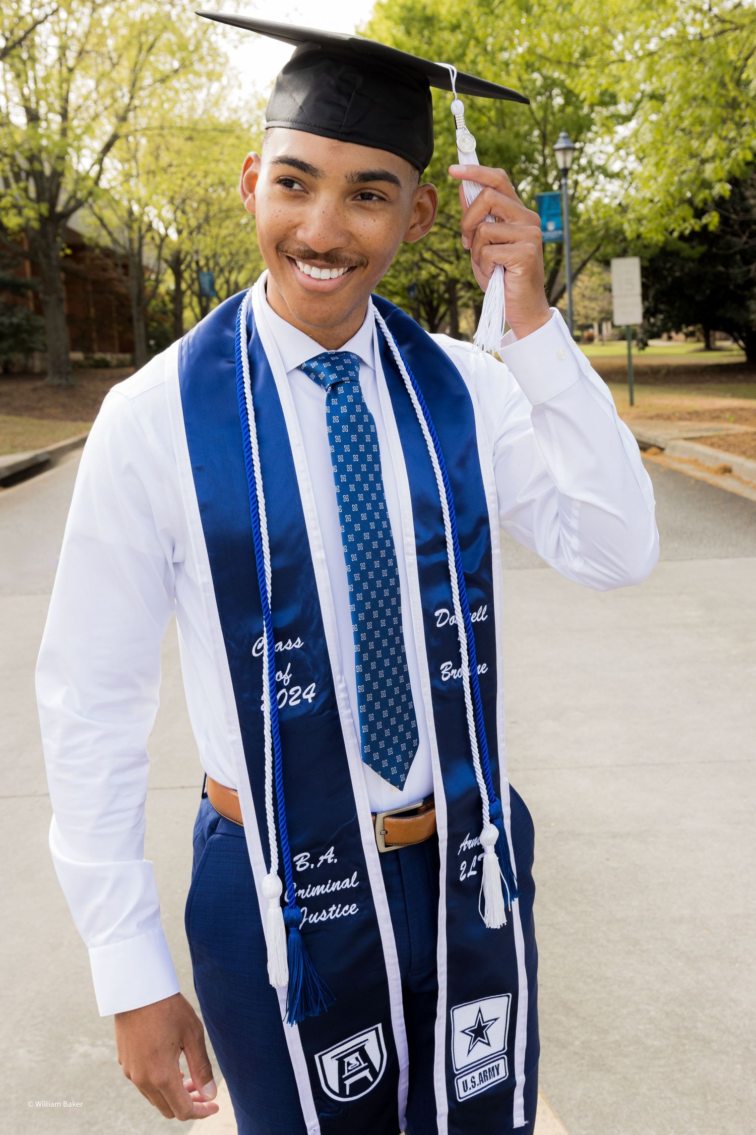Graduate wearing white academic regalia with blue and white stole and polka dot tie at outdoor ceremony.