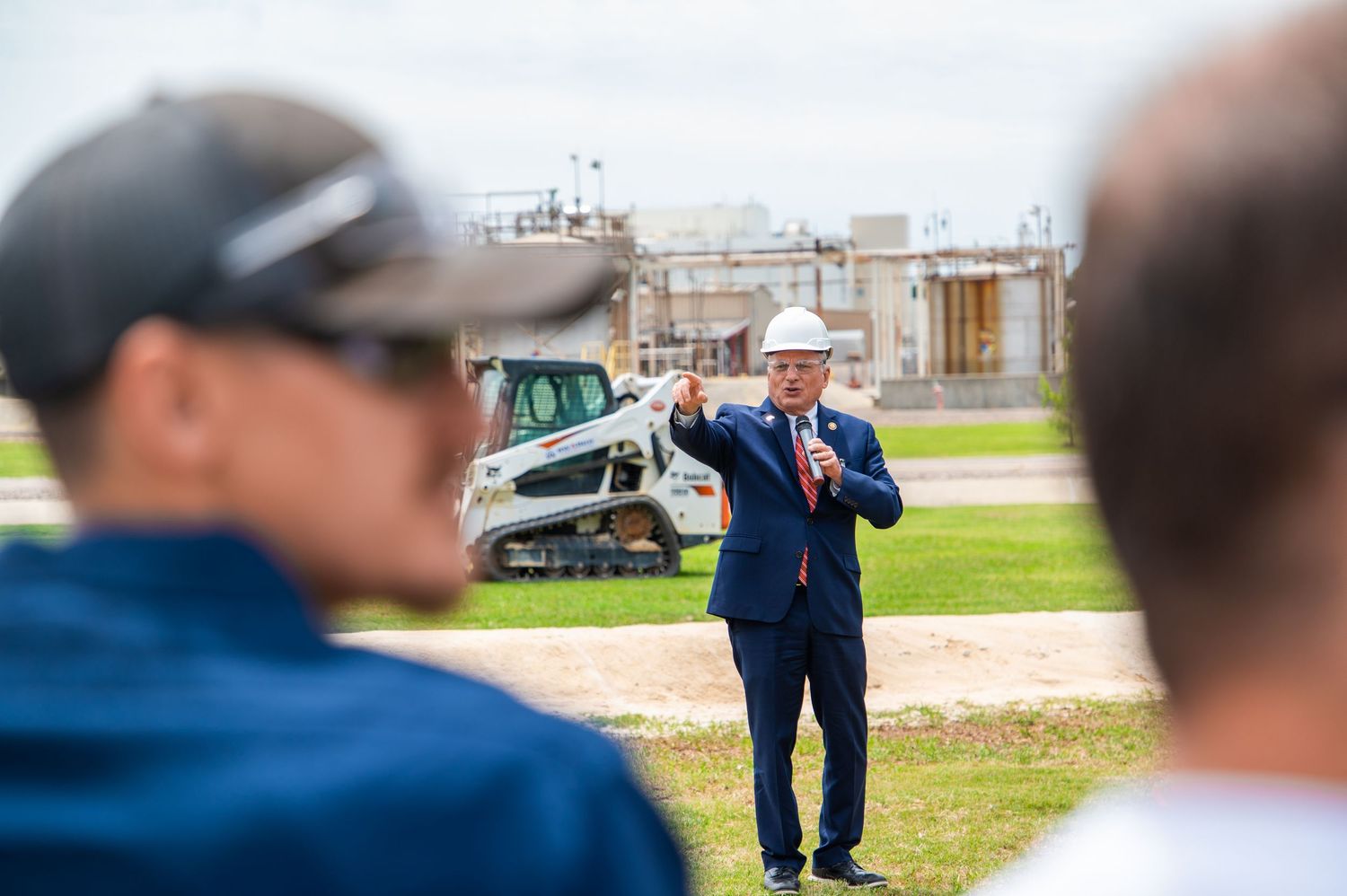 A man in a white hard hat and suit speaks to a crowd at an industrial construction site with heavy machinery in the background.