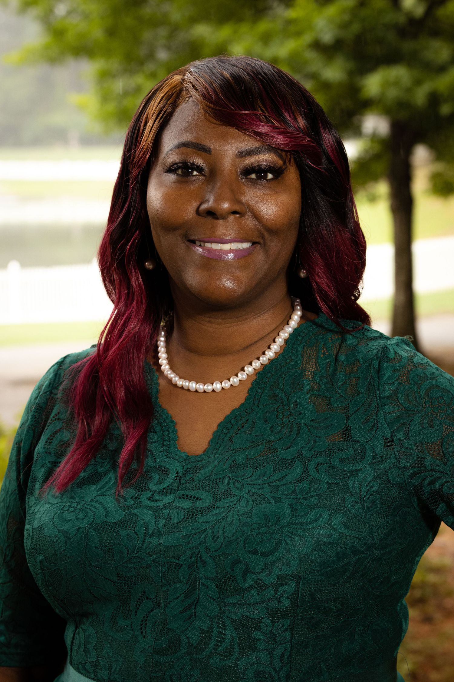 Woman in green lace dress and pearl necklace with red highlighted hair smiling outdoors in a park setting.