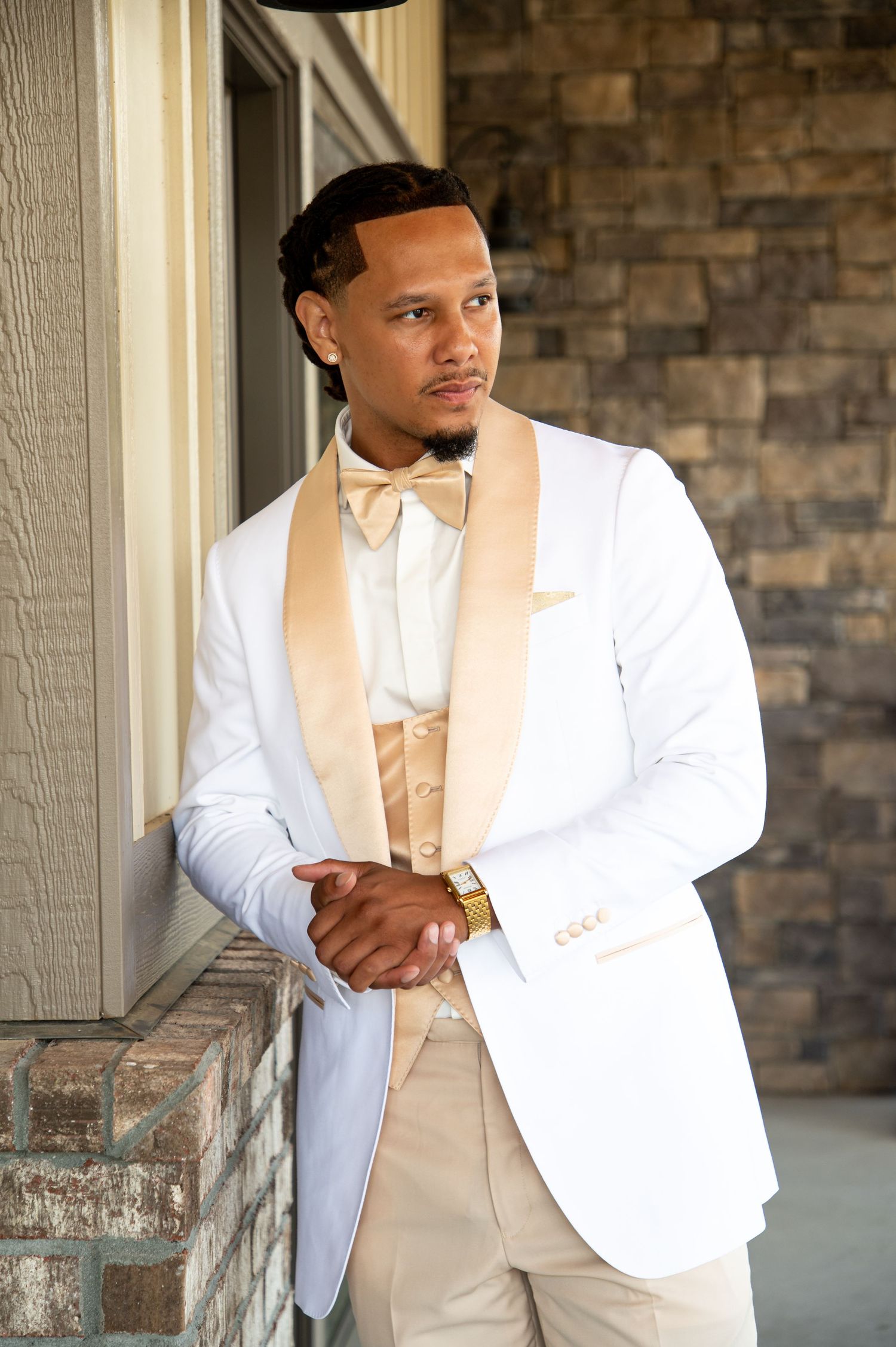 Stylish groom in white and champagne tuxedo with gold watch posing against stone wall at wedding.