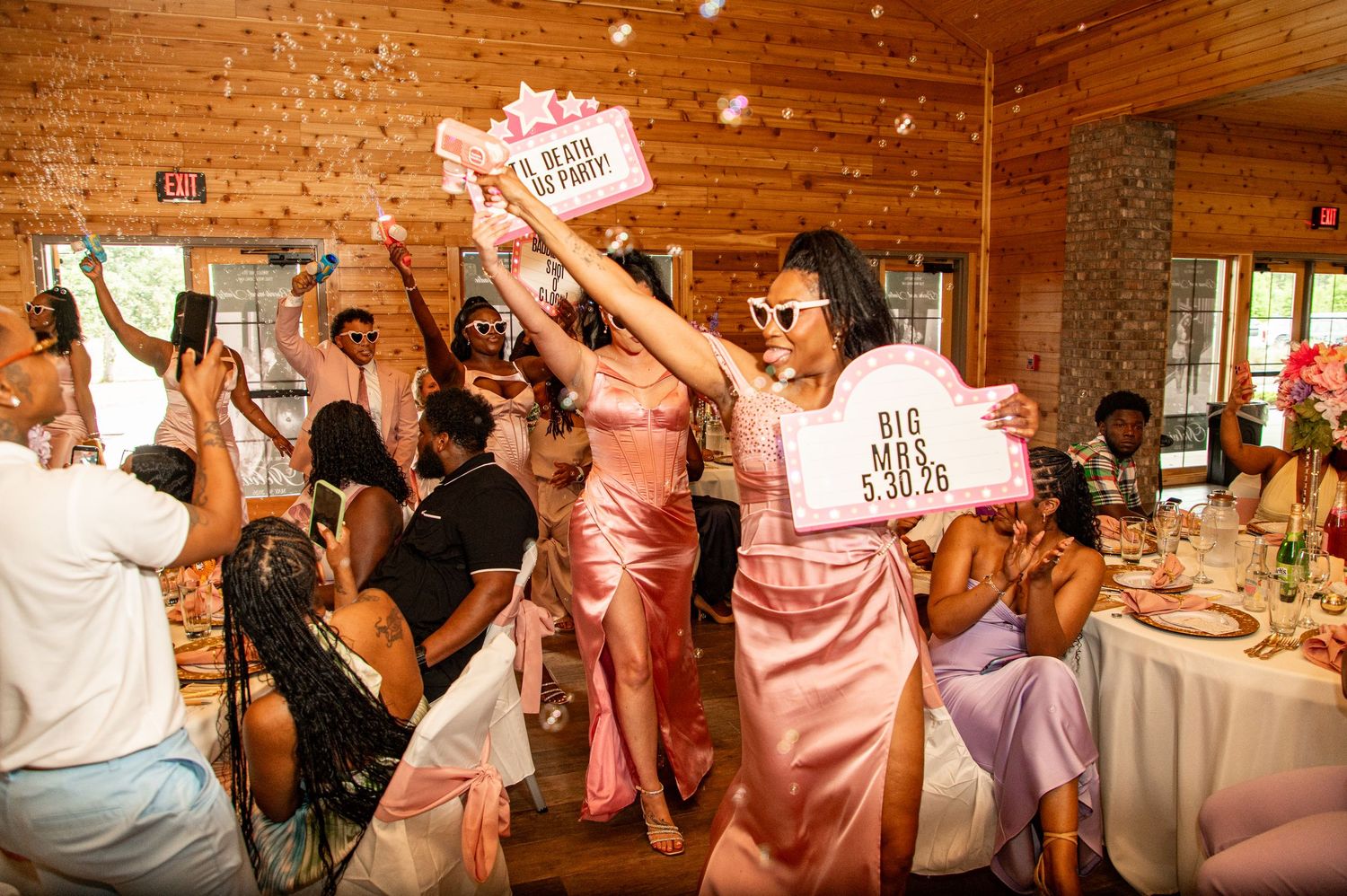 Bridesmaids in pink dresses celebrate at lively wedding reception holding decorative signs.