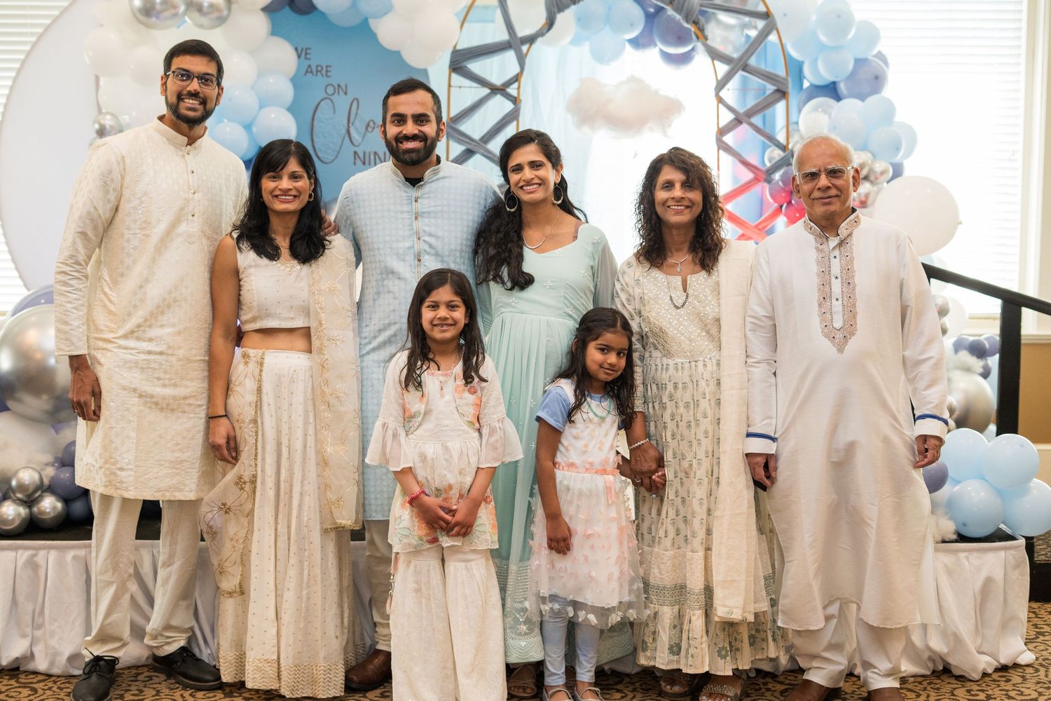 A group in traditional cream and white Indian ethnic wear poses together against a backdrop with light blue balloons.