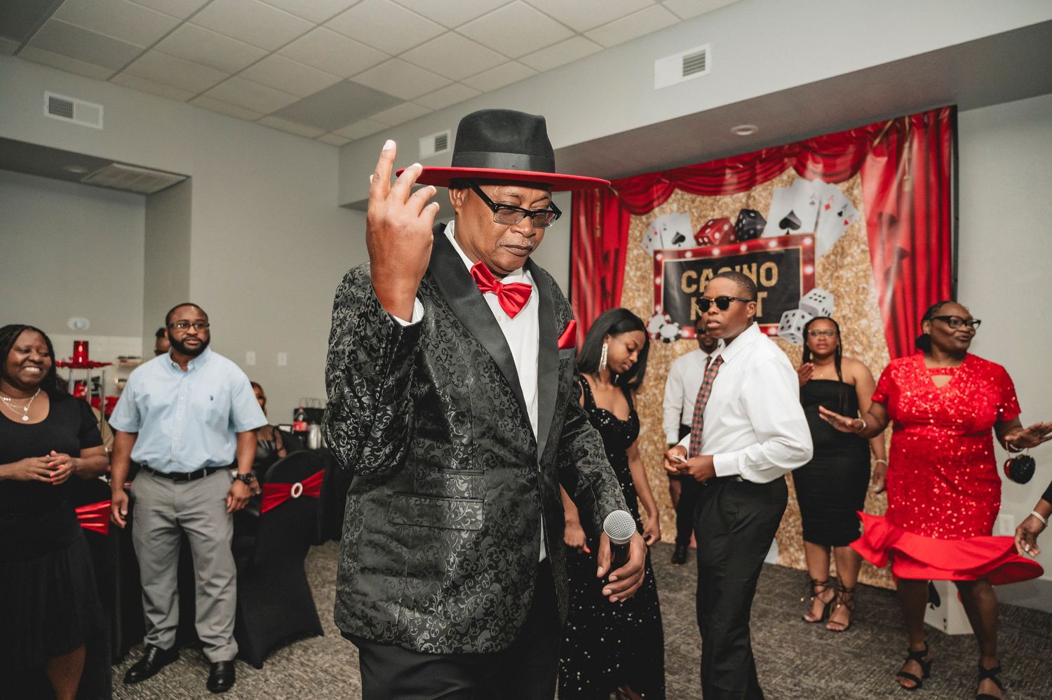 Group of party guests dancing and socializing at a casino themed celebration with red curtain backdrop.