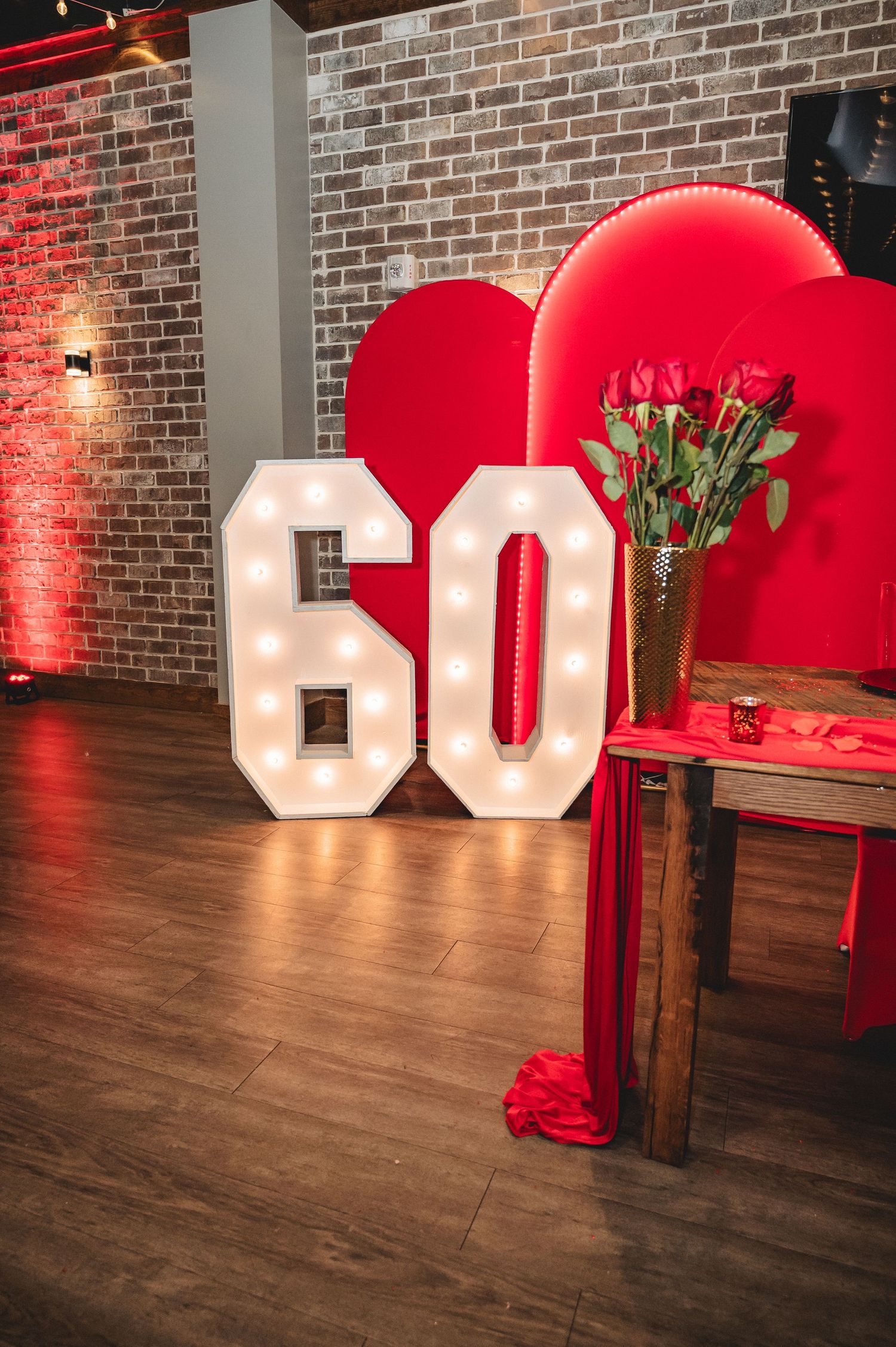Large illuminated '60' numbers decorate a rustic brick wall venue with red accents and wooden floors.