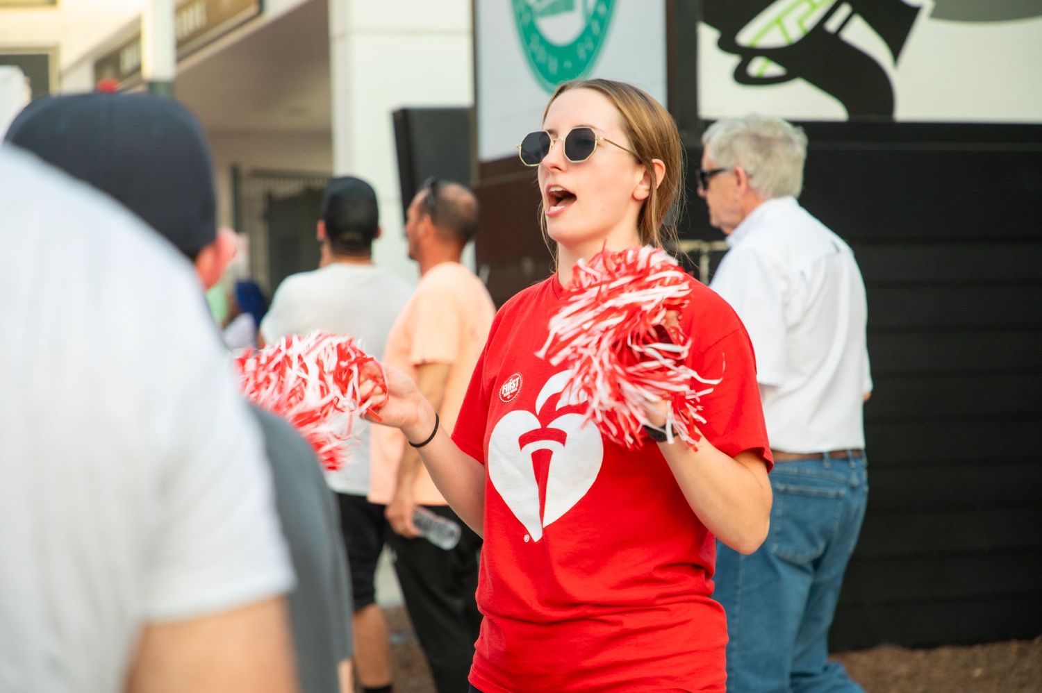 Woman in red American Heart Association t-shirt cheering with red and white pom-poms at an outdoor event.