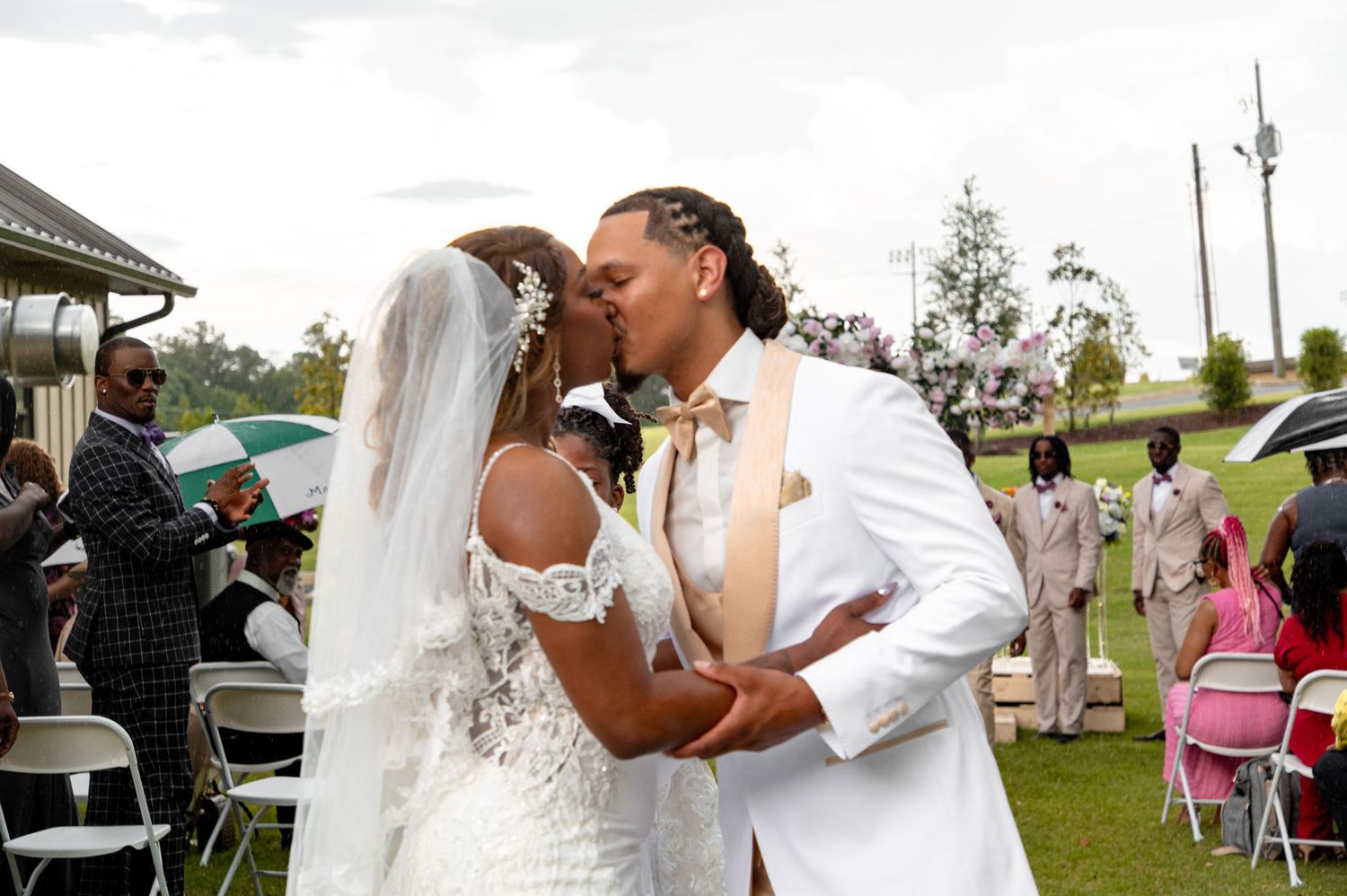 Newlyweds share first kiss at outdoor wedding ceremony surrounded by guests.