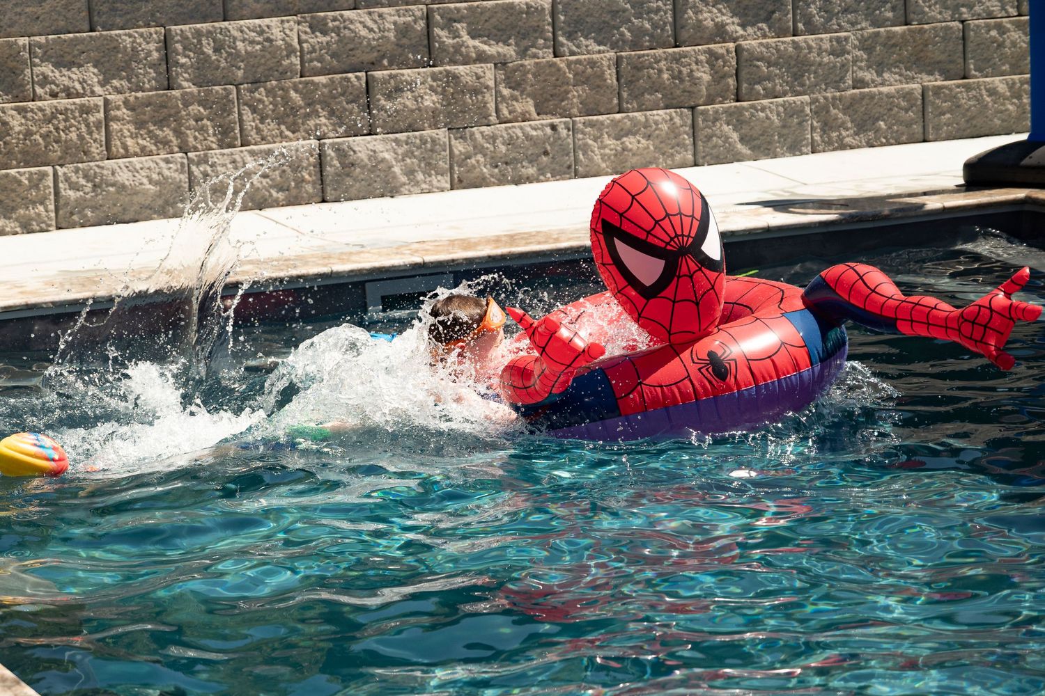 Person in Spider-Man costume swimming in a pool with an inflatable ring, splashing water on a sunny day.