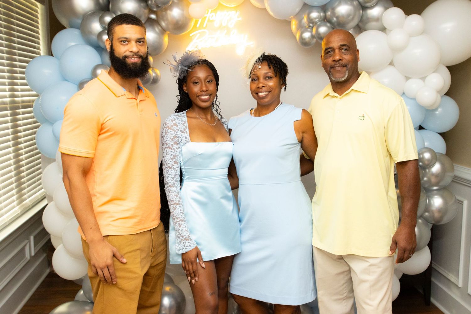 Four people in coordinated pastel outfits pose together in front of a decorative balloon arch at a celebration.