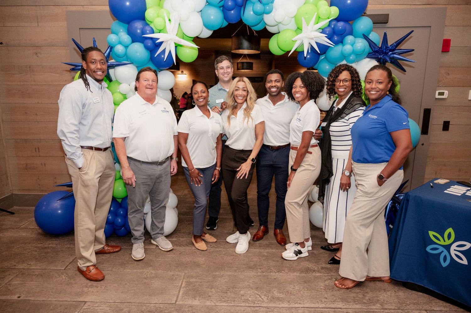 A group of people in white and blue attire pose together at an event with blue and green balloon decorations.