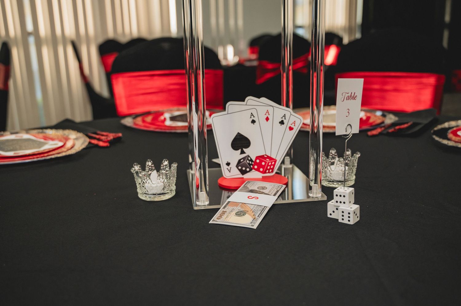 Casino themed party decorations including playing cards, dice, crown decor, and money on a black tablecloth with red accents.