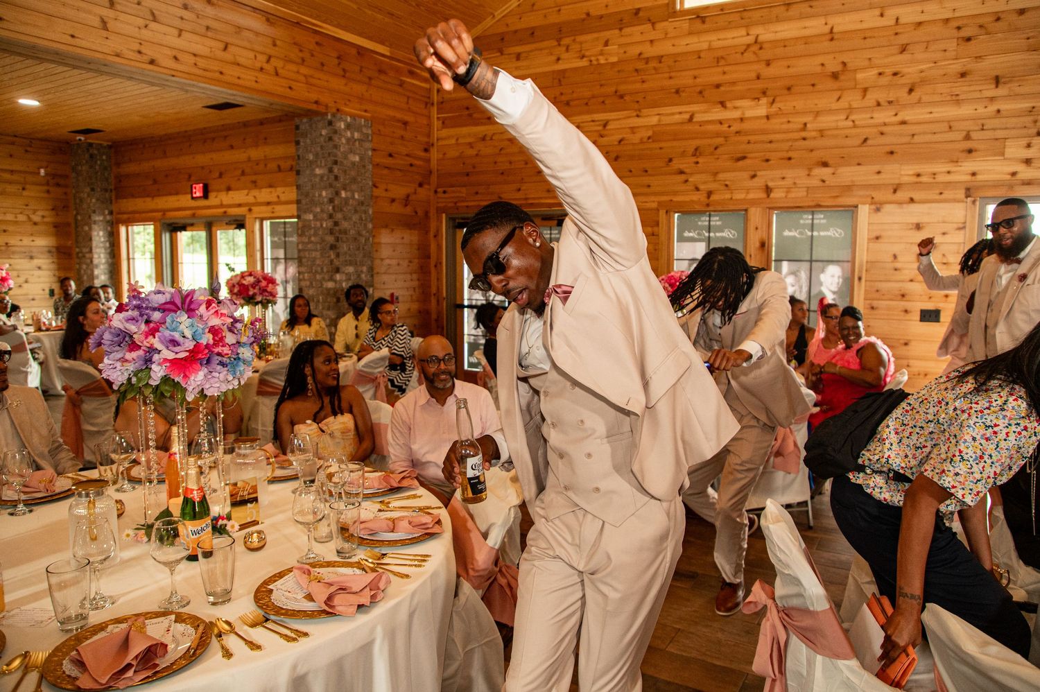 Groom in white suit raises hand joyfully while dancing at wedding reception in rustic log cabin venue.