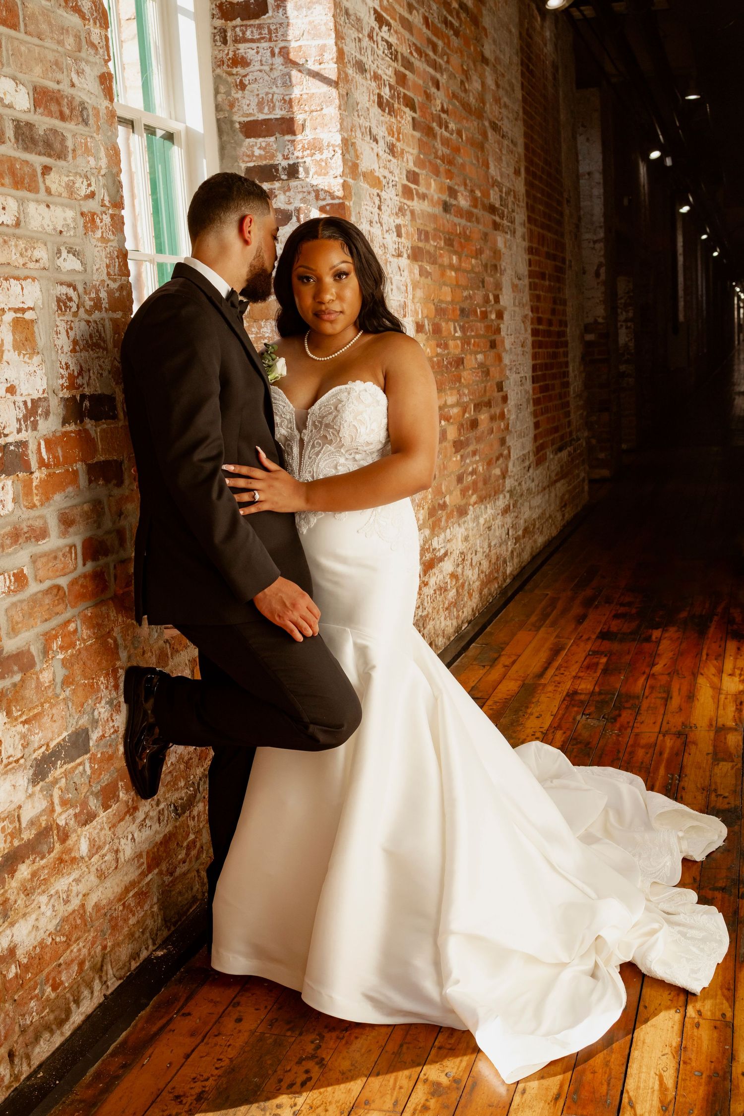 Bride in white mermaid gown and groom in black suit pose romantically against rustic brick wall on hardwood floors.