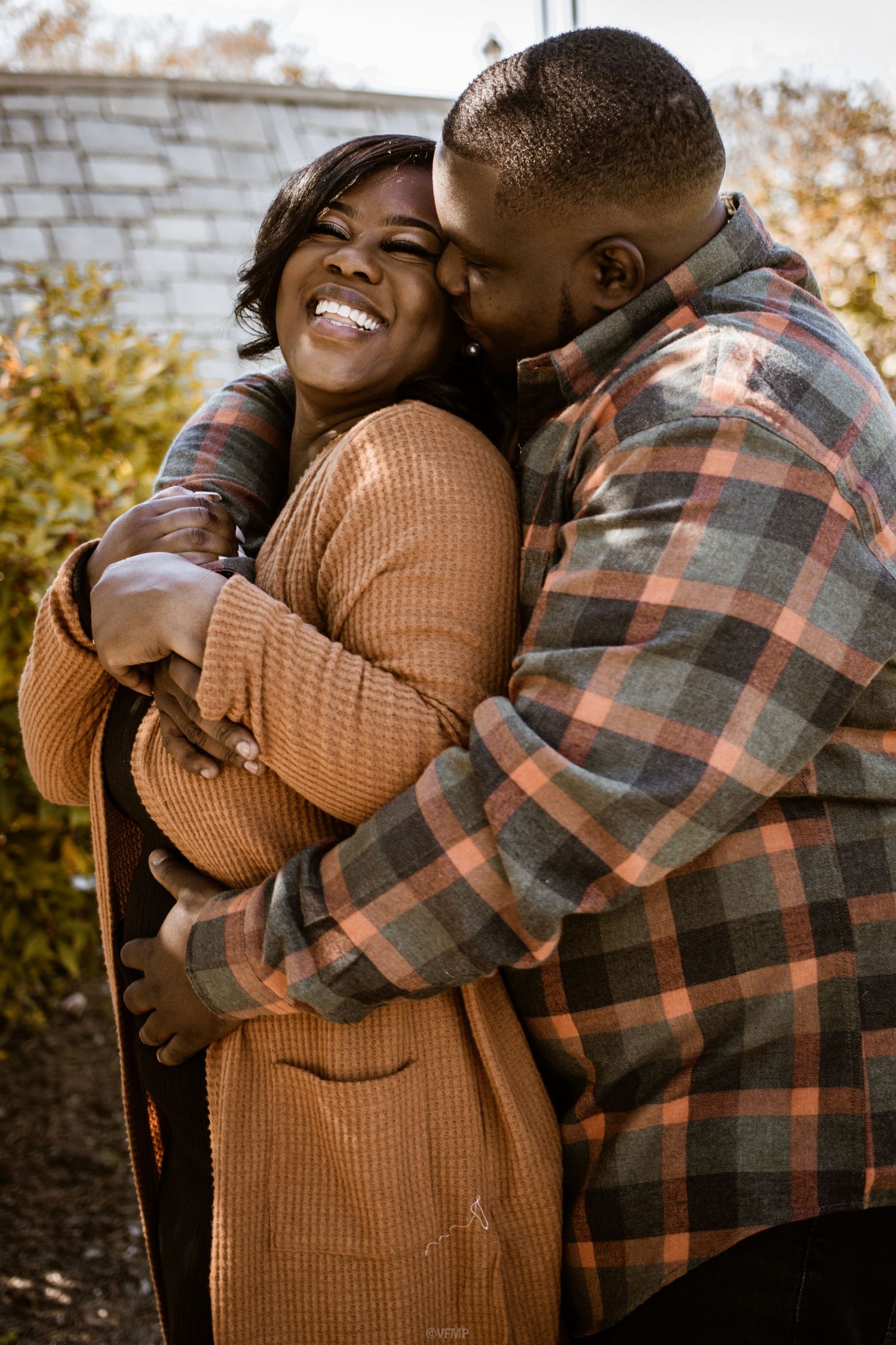 A couple shares an intimate embrace outdoors, wearing fall clothing and expressing joy and affection.