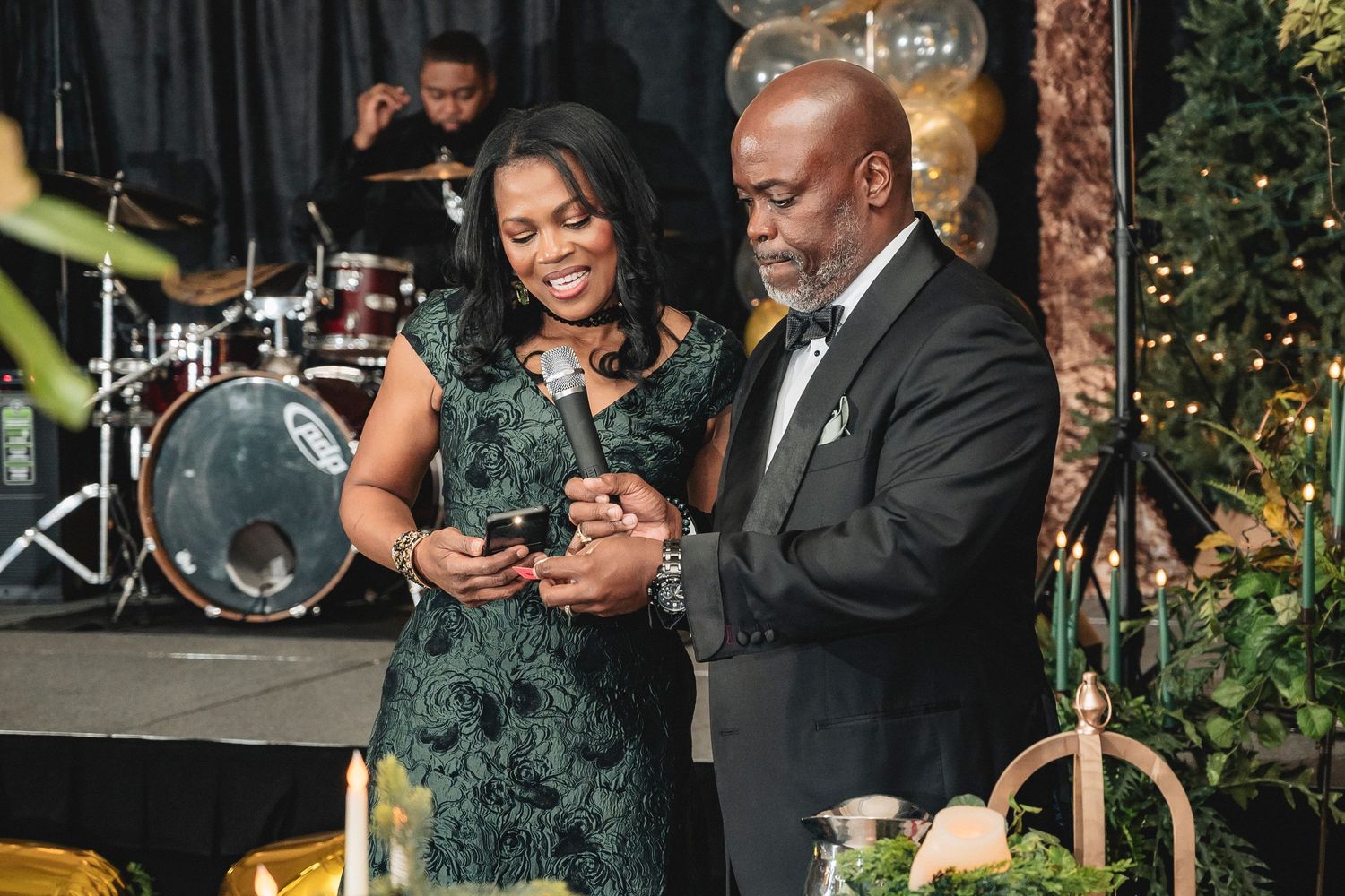 Two people in formal attire look at a phone together at an elegant outdoor event with musical instruments in background.