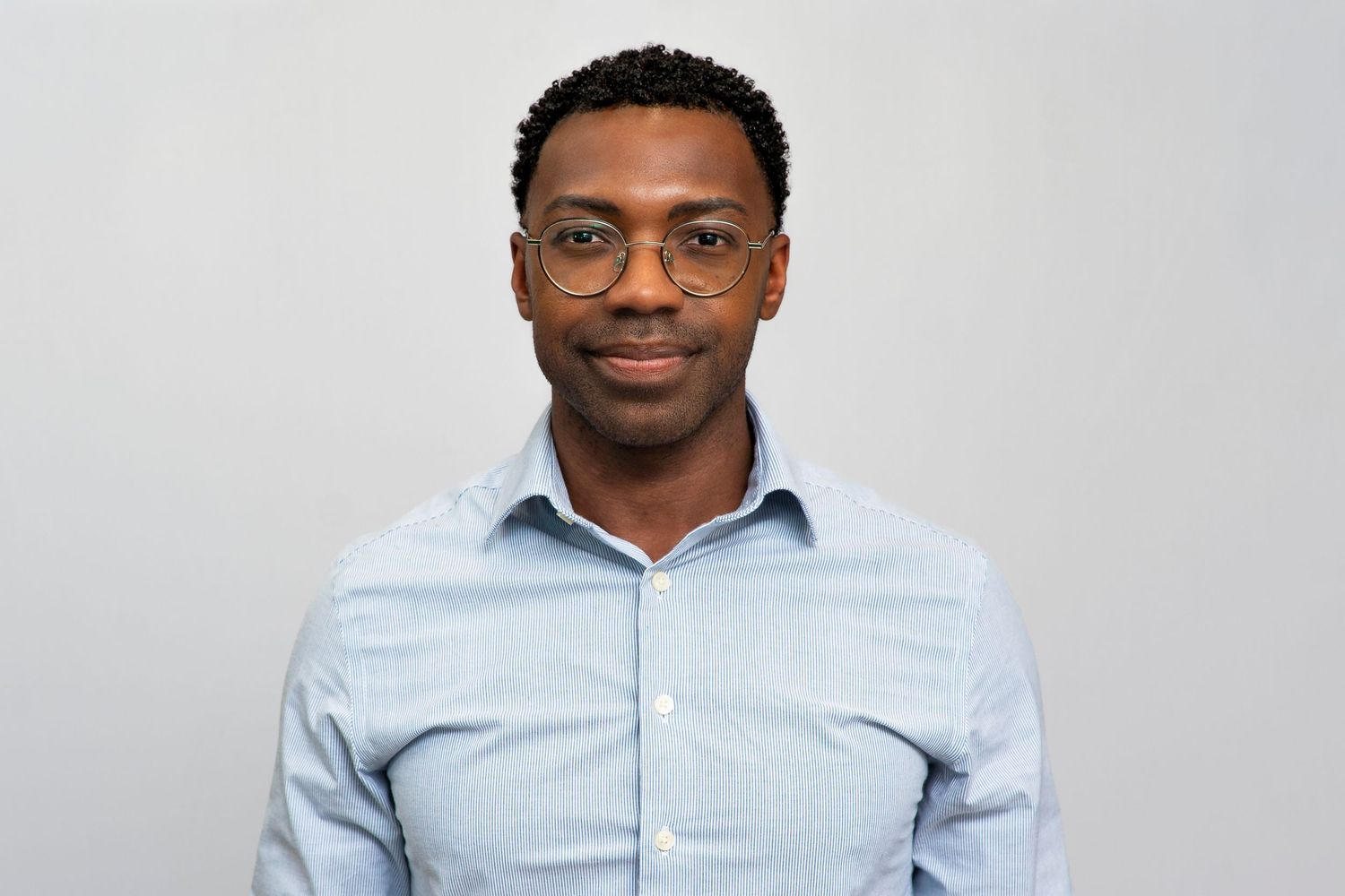 Young professional man wearing glasses and a light blue striped shirt, smiling confidently against a gray background.