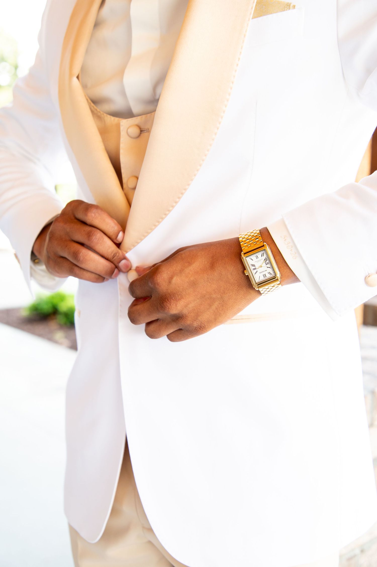 Groom in white tuxedo jacket with gold lapels fastening button, wearing gold watch.