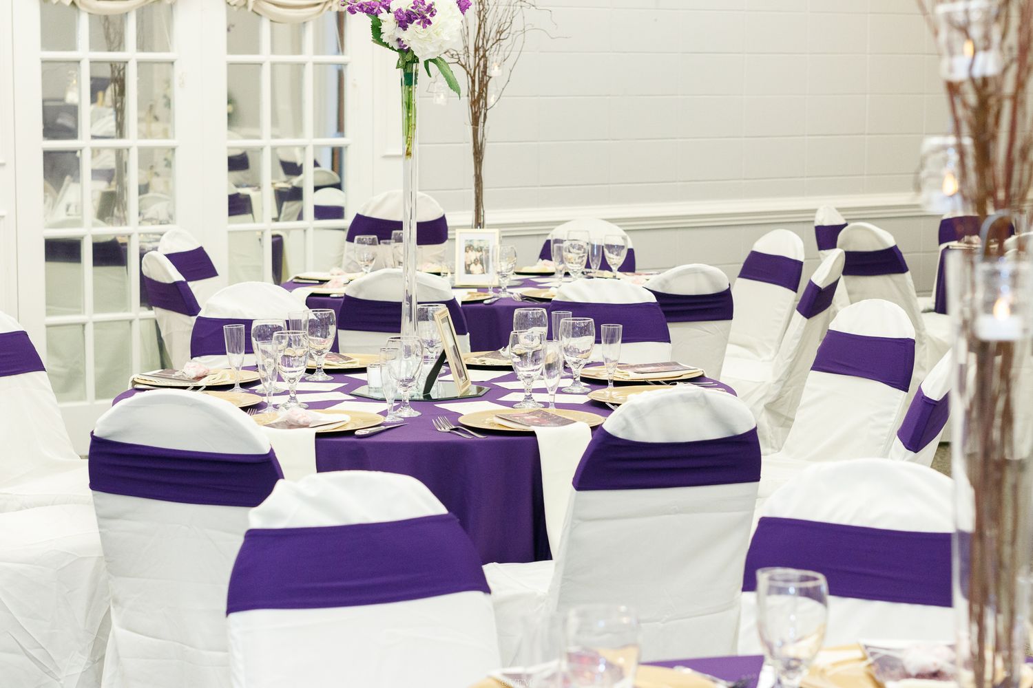 Elegant wedding reception tables with white linens and royal purple chair sashes in a bright sunlit venue.