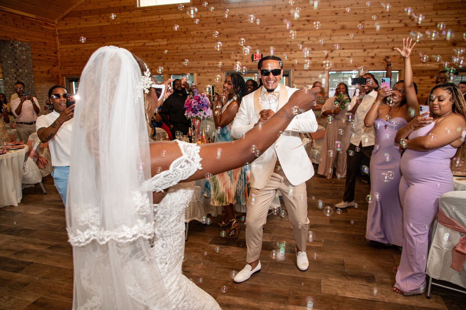 Joyful bride and groom sharing first dance surrounded by cheering guests and bubbles at rustic wedding reception.