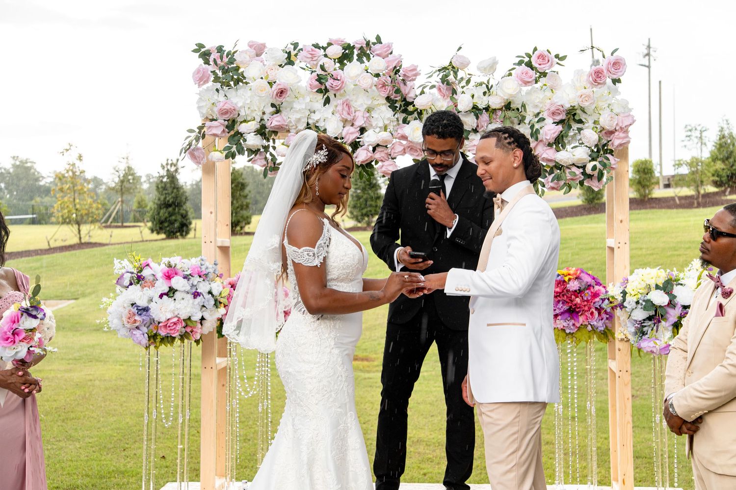 Bride and groom exchanging rings at outdoor ceremony beneath floral arch.