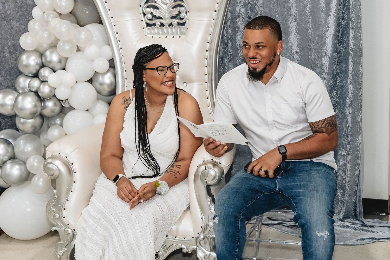 A couple sits together on a white throne chair surrounded by silver and white balloon decorations.