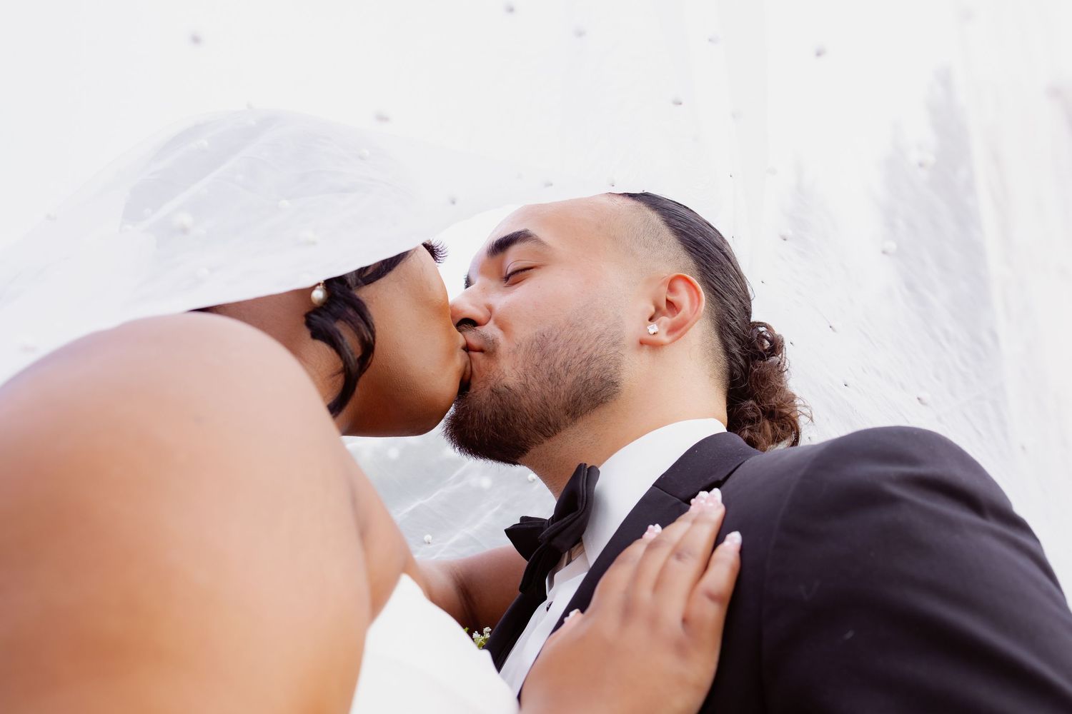 A couple shares a romantic kiss outdoors with a bright sky in the background.