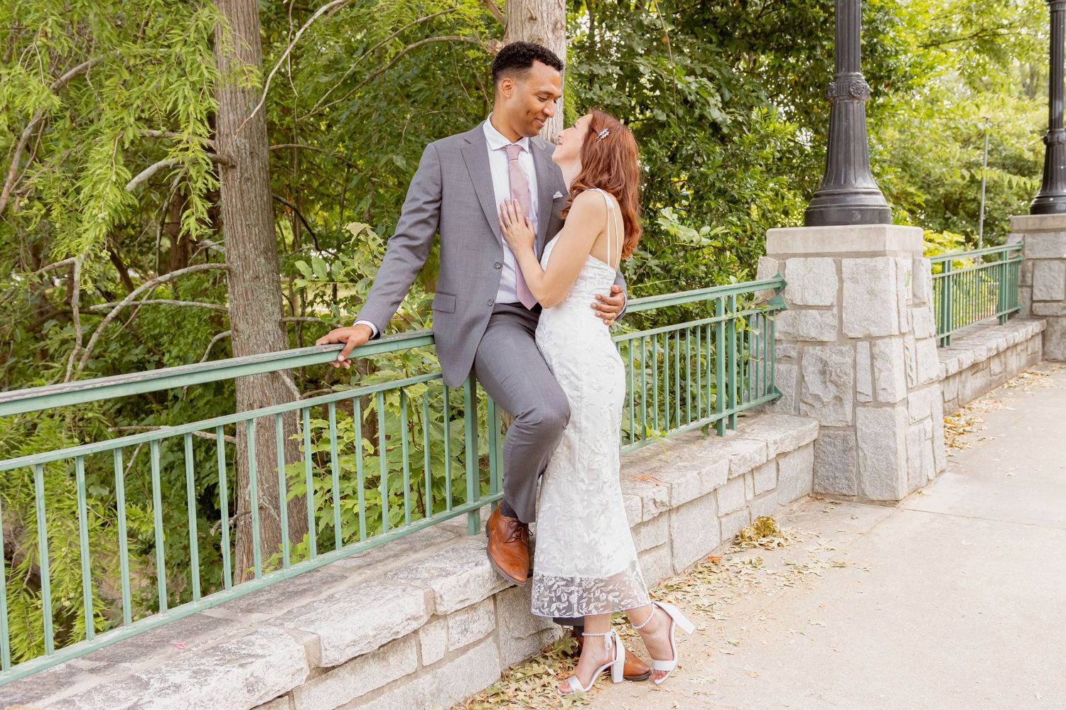 A couple in wedding attire shares a romantic moment by a green railing surrounded by lush trees.