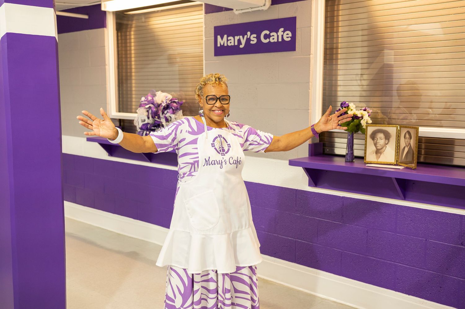 Woman in purple and white outfit stands proudly in front of Mary's Cafe counter with arms outstretched.