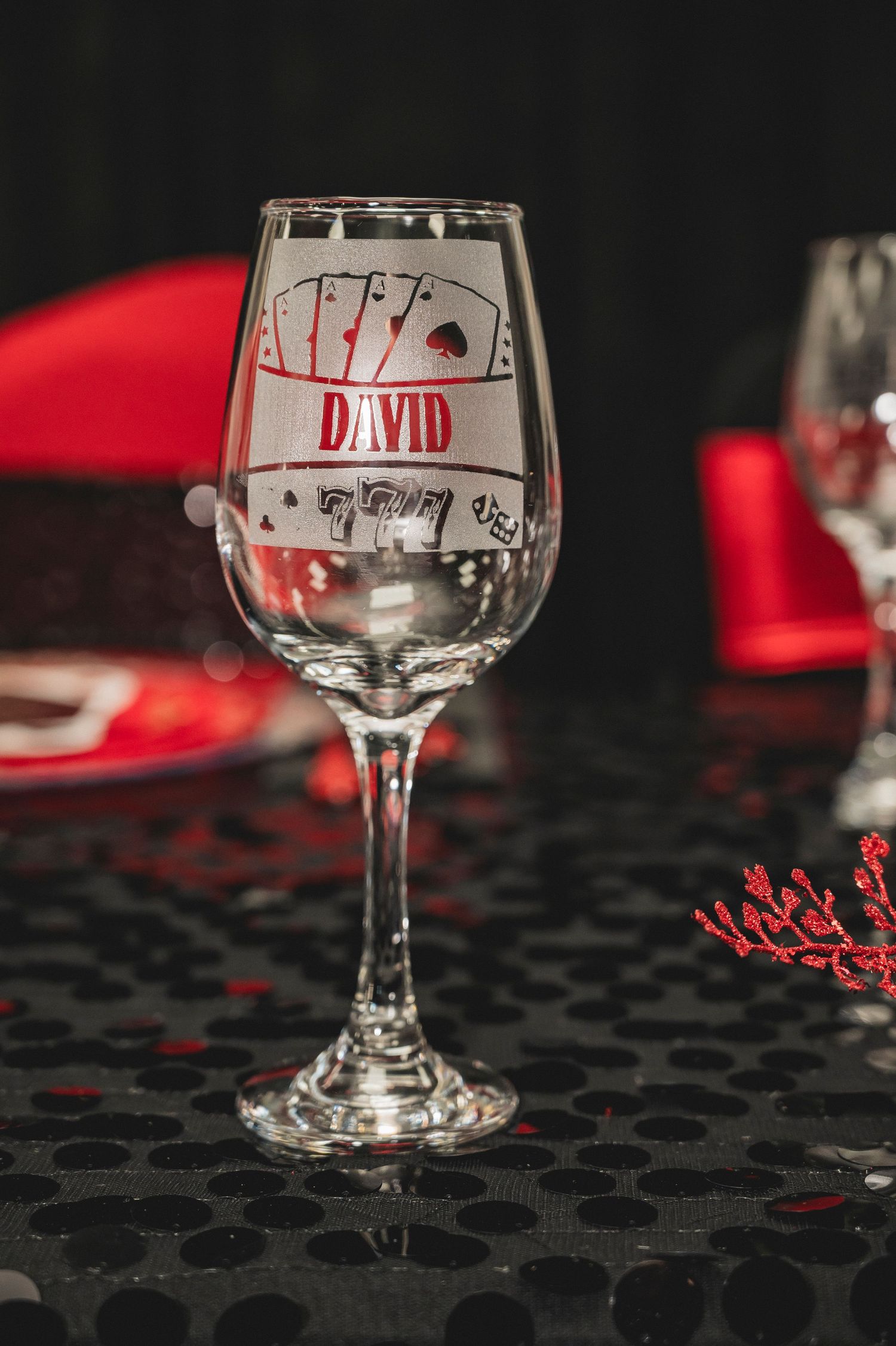Wine glass with personalized etching sits on dark surface with red accents and decorative elements.