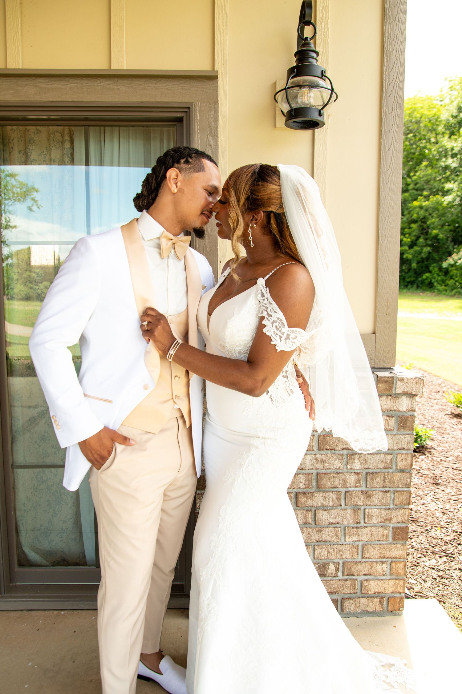 A bride and groom in elegant white and champagne wedding attire share a romantic moment outdoors near a brick building.