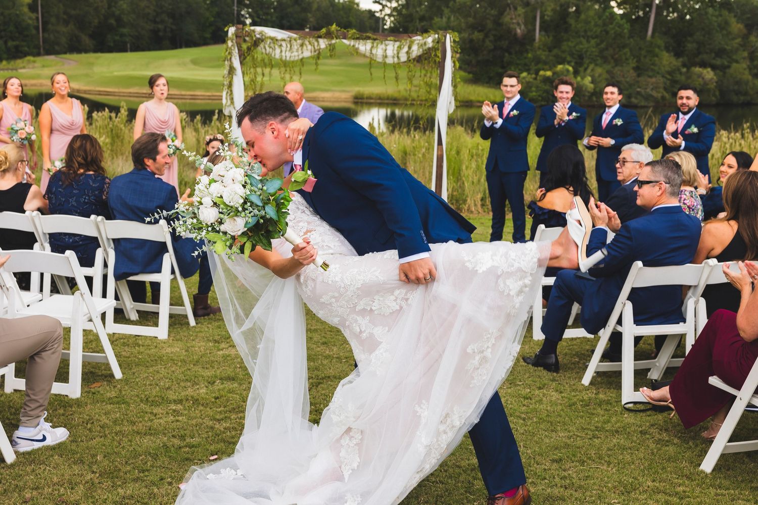 A wedding ceremony setup in an outdoor garden with white chairs and floral decorations along a grassy aisle.