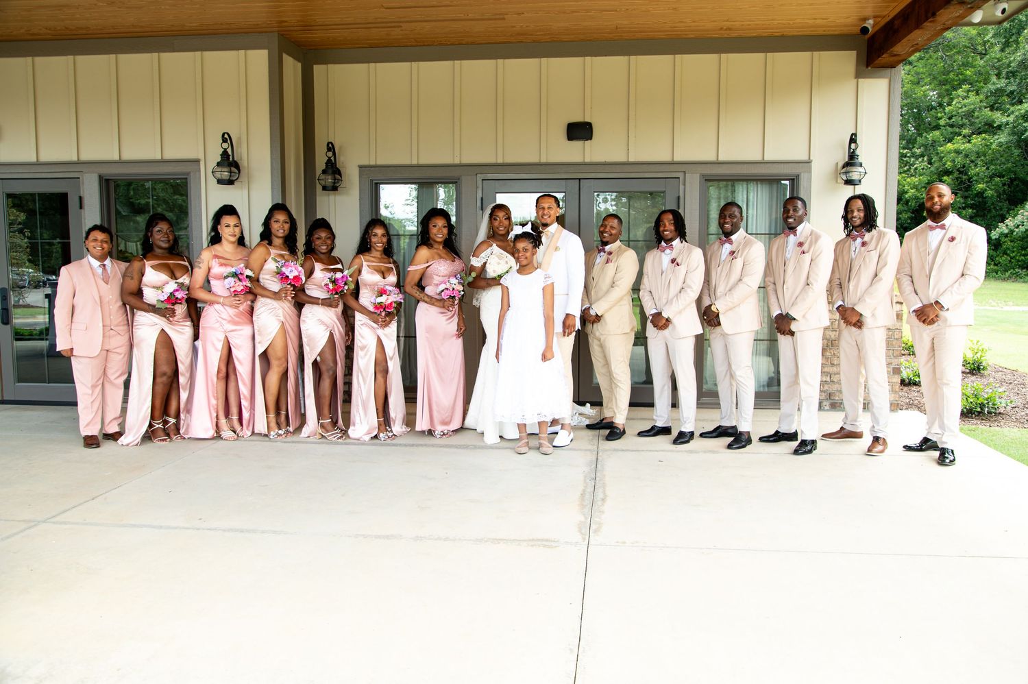 Wedding party in pink and beige attire posing under venue pavilion.