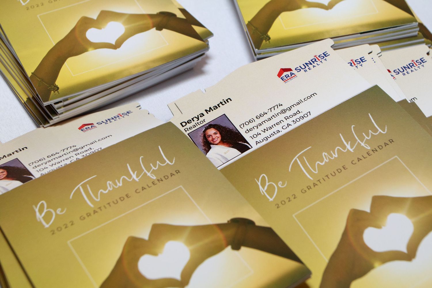 Stack of 'Be Thankful 2022 Gratitude Calendar' booklets with a heart hands silhouette cover design by Sunrise Realty.