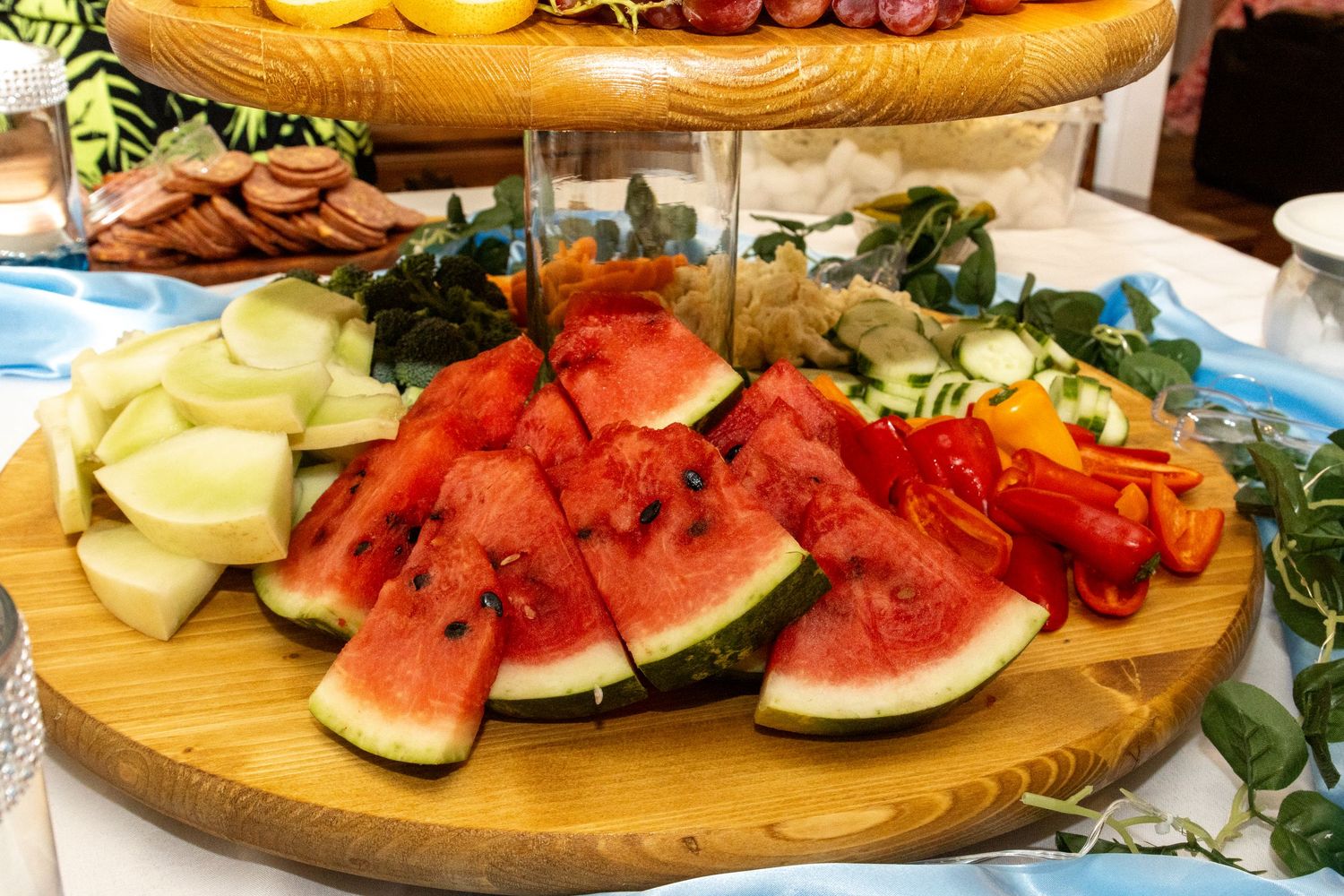 Fresh watermelon slices and other fruits arranged on a wooden circular serving platter at a party buffet.