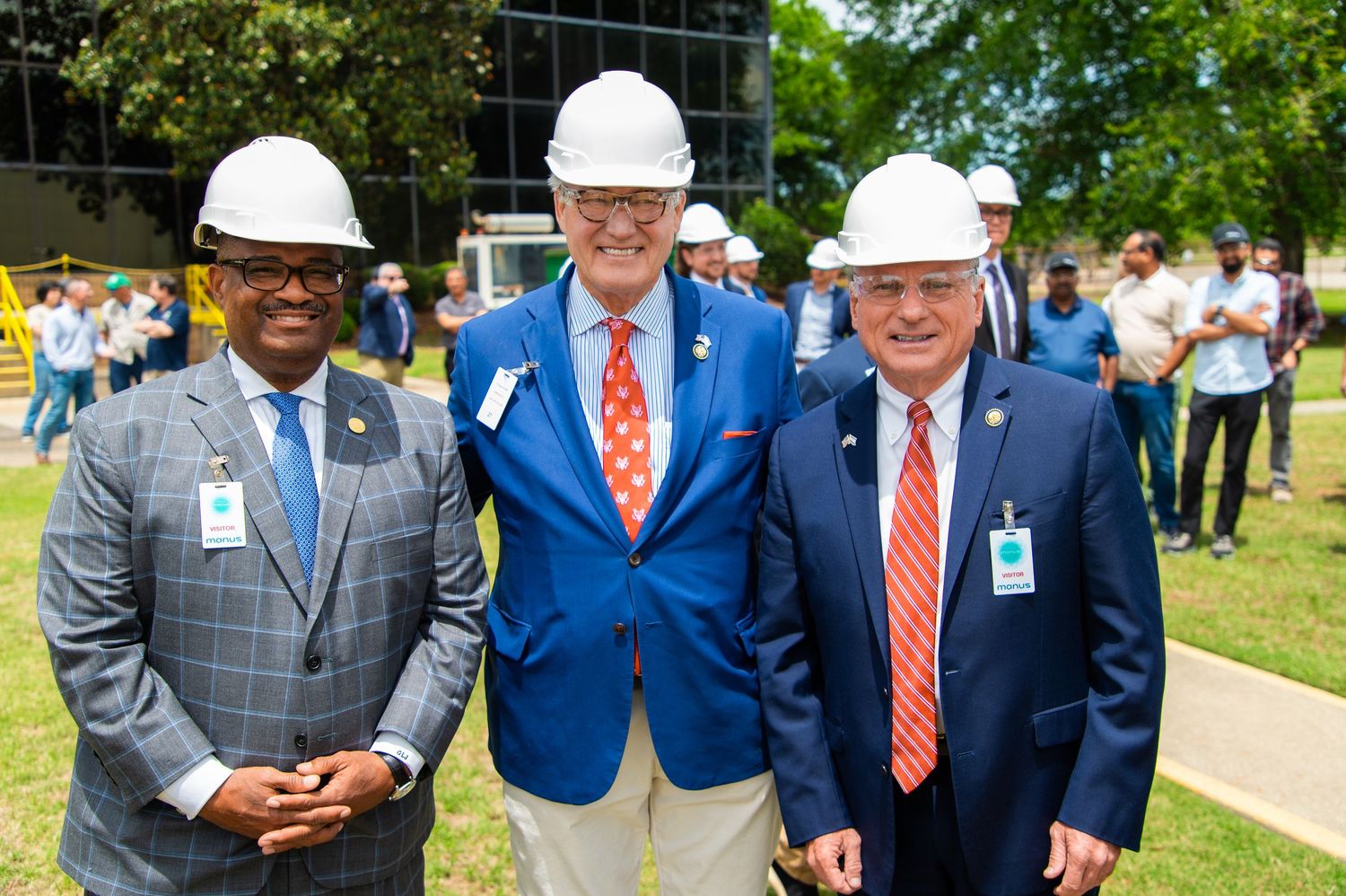 Three men in hard hats and suits smile outdoors at a construction groundbreaking ceremony.