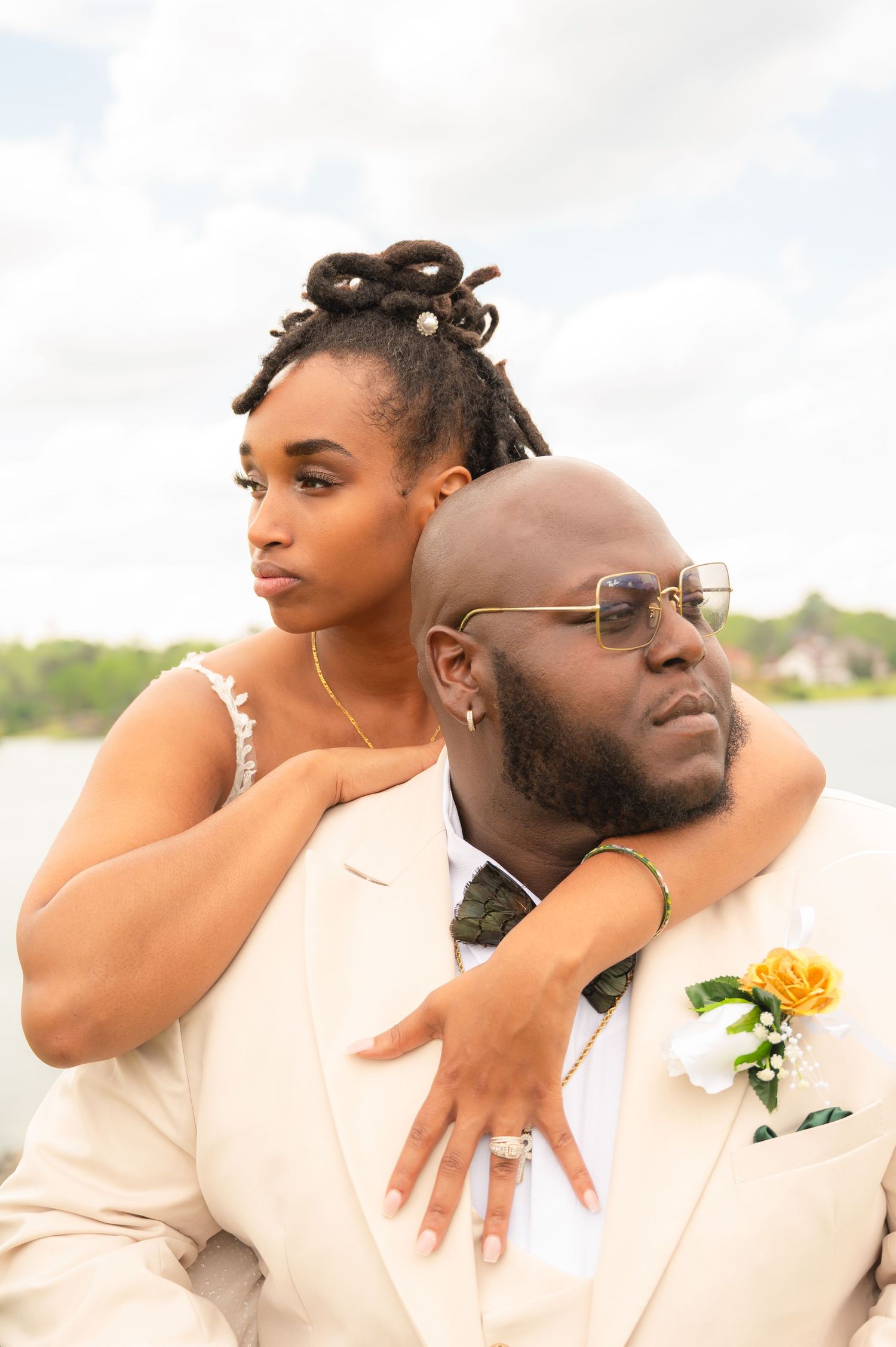 A bride and groom in cream attire pose outdoors near water, she embraces him from behind with a yellow boutonniere visible.