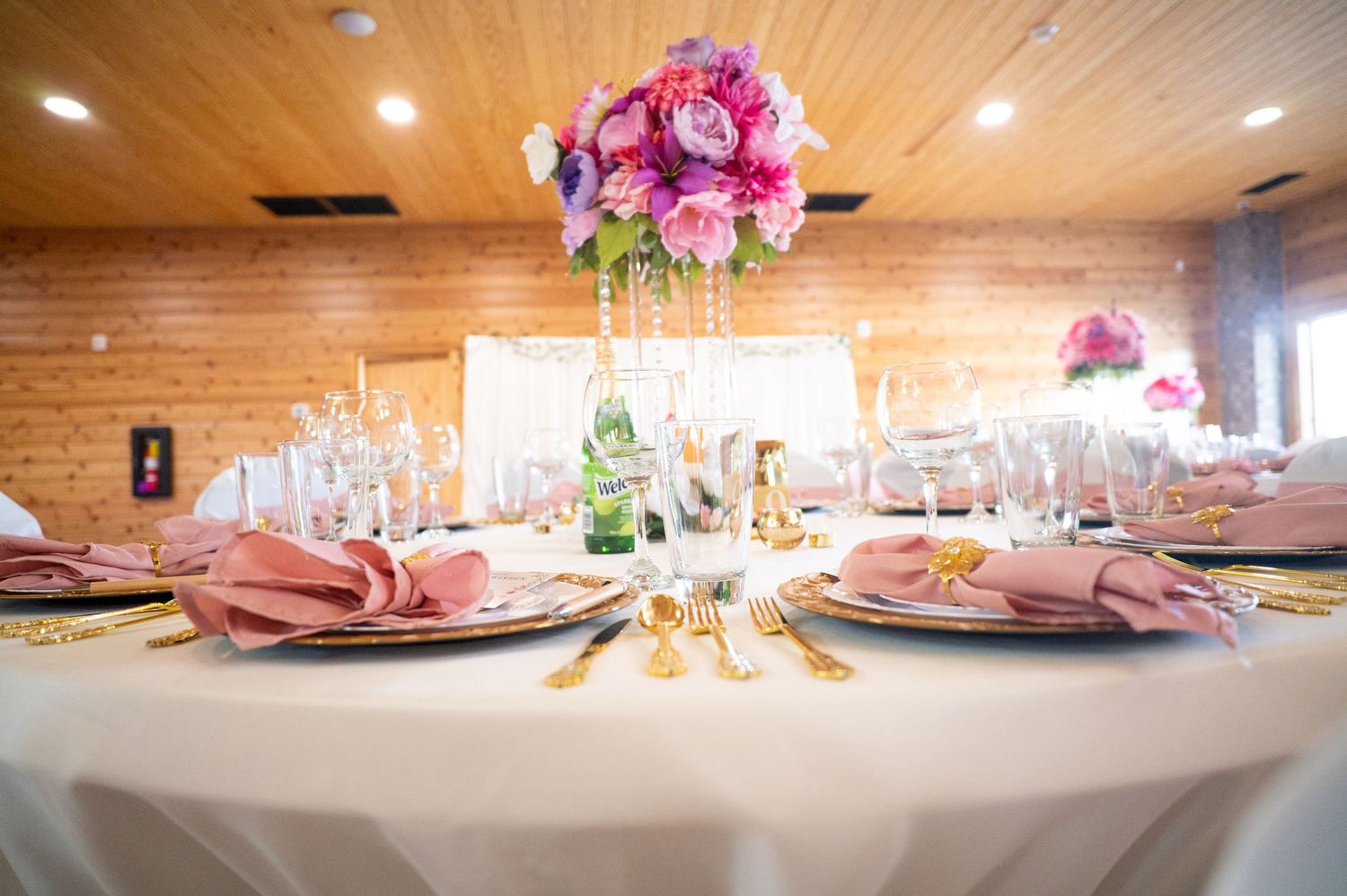 Elegant wedding reception table with pink floral centerpiece, gold cutlery, and dusty rose napkins in a rustic venue.