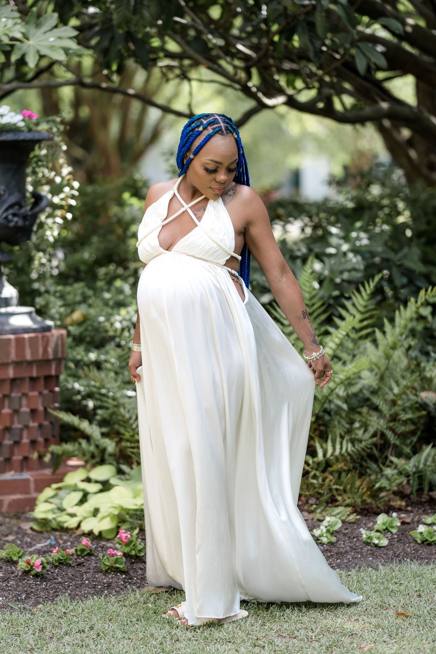 A person in a flowing white dress kneels gracefully in a garden setting with lush greenery and brick walls.