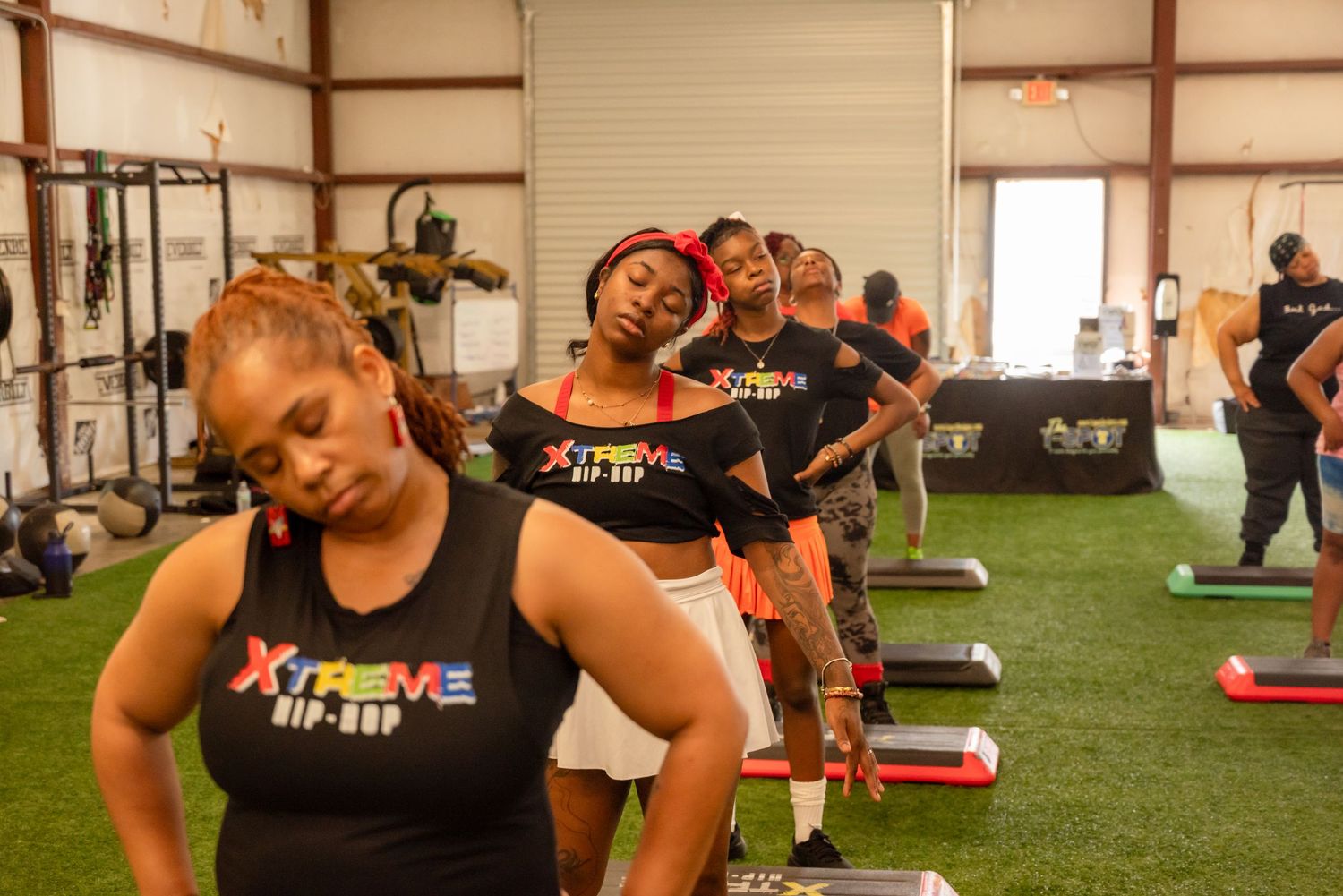 A group works out in a fitness facility on red floor mats with exercise equipment visible in the background.