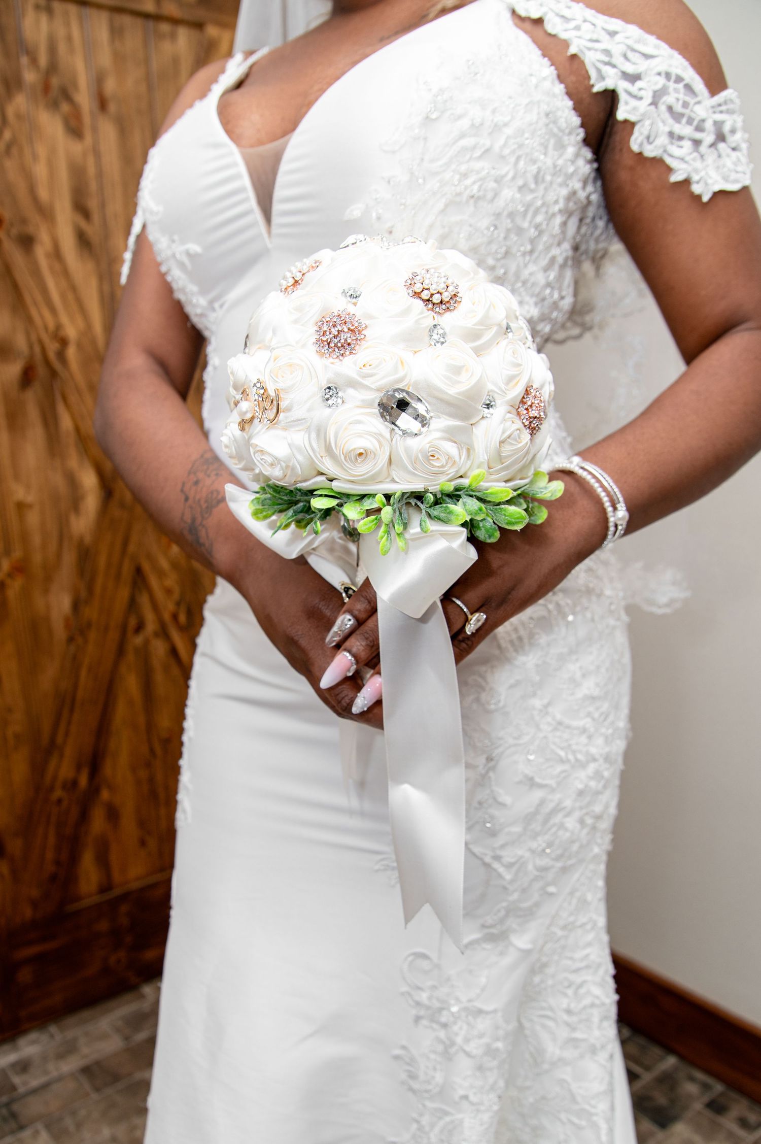 Bride in lace wedding gown holds white and rose gold brooch bouquet with ribbon.
