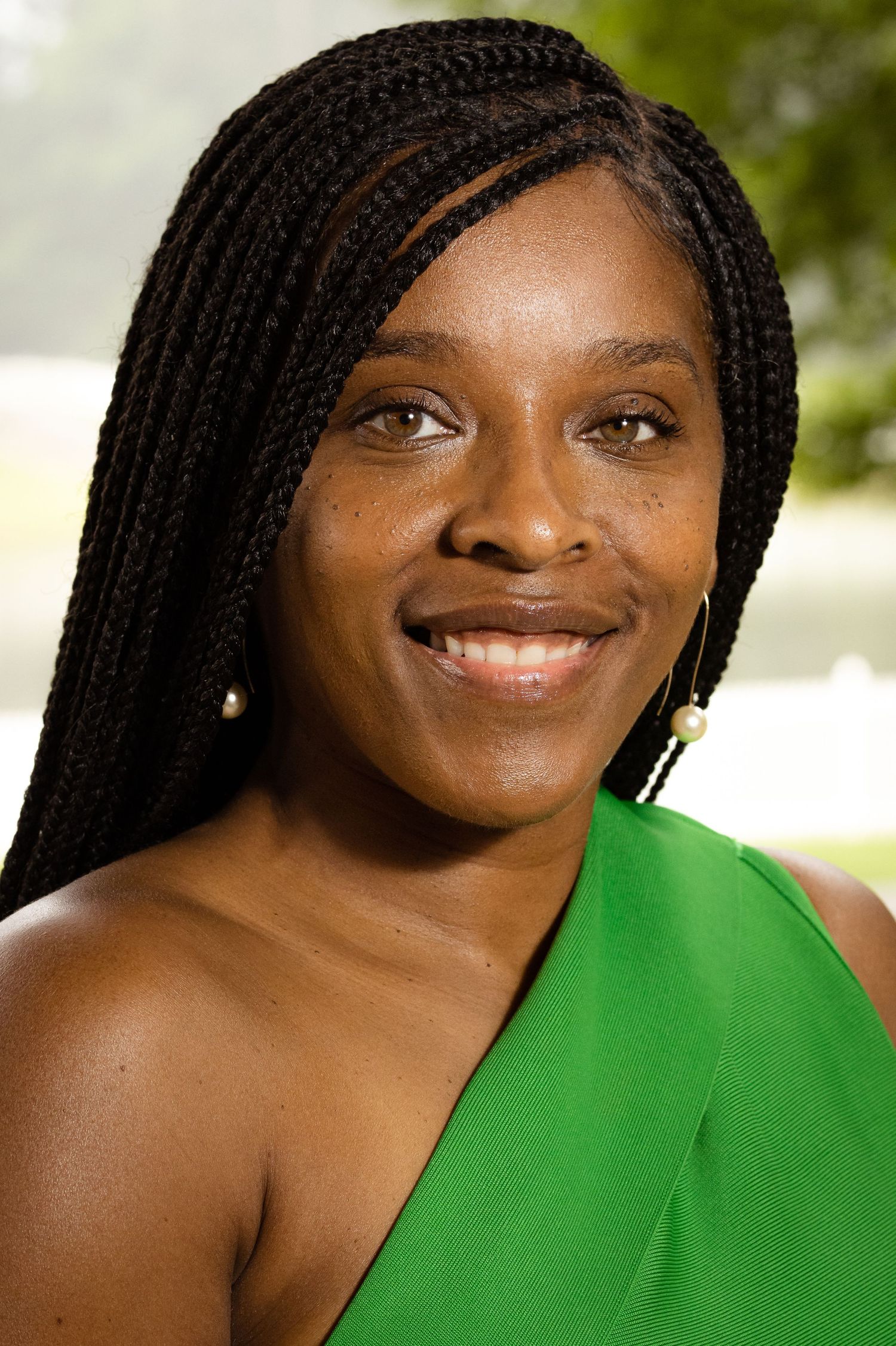 Smiling woman with braids wearing a green one-shoulder top outdoors.