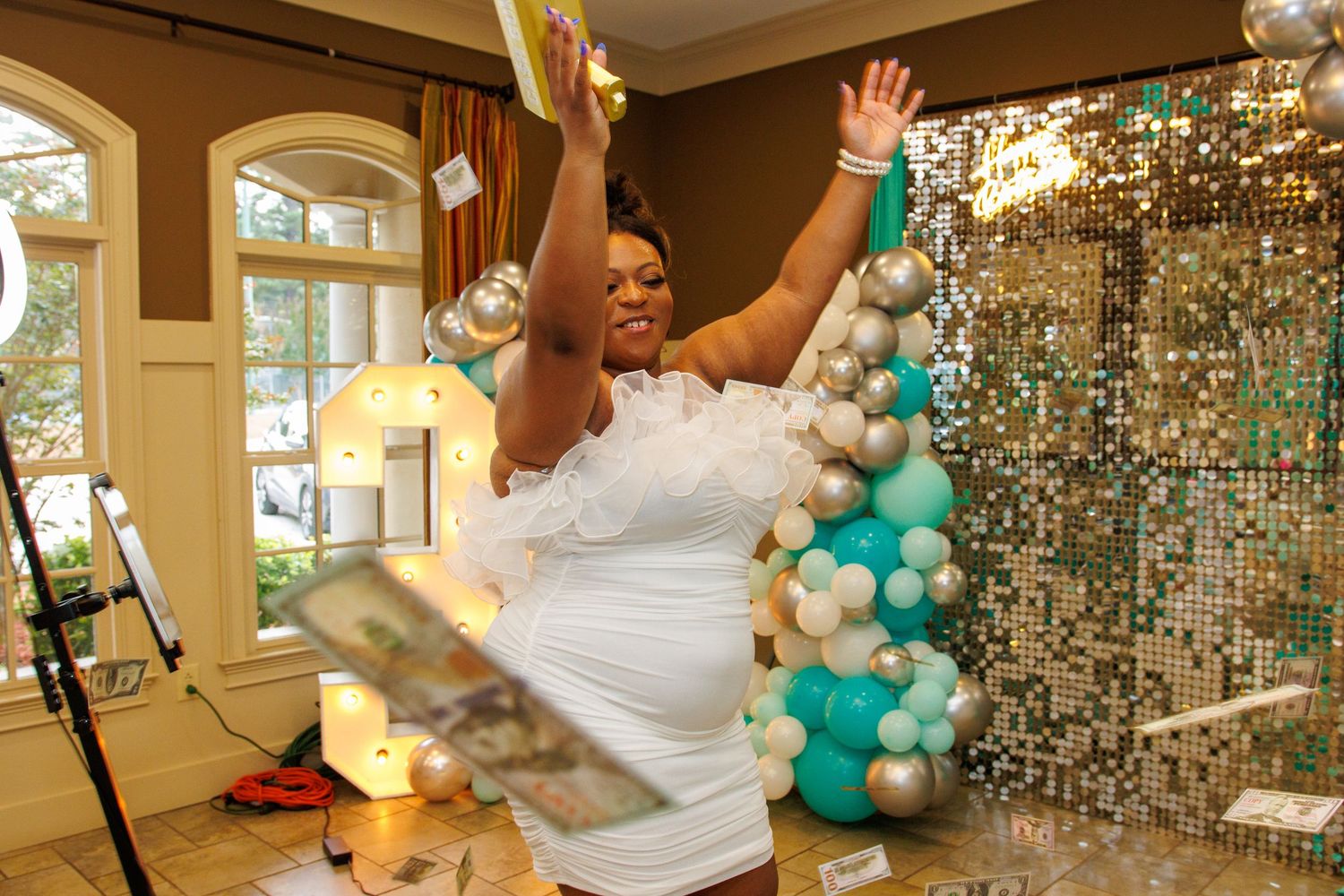 Woman in white dress celebrating with arms raised at festive party with balloon arch and gold sequin backdrop.